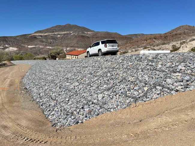 Gray rocks in wire baskets compose a large flood-protection berm. A historic building with a red roof can be seen in the distance.