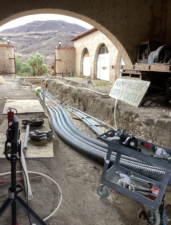 Gray pipes in a dirt trench surrounded by tan-colored historic building with a red roof.