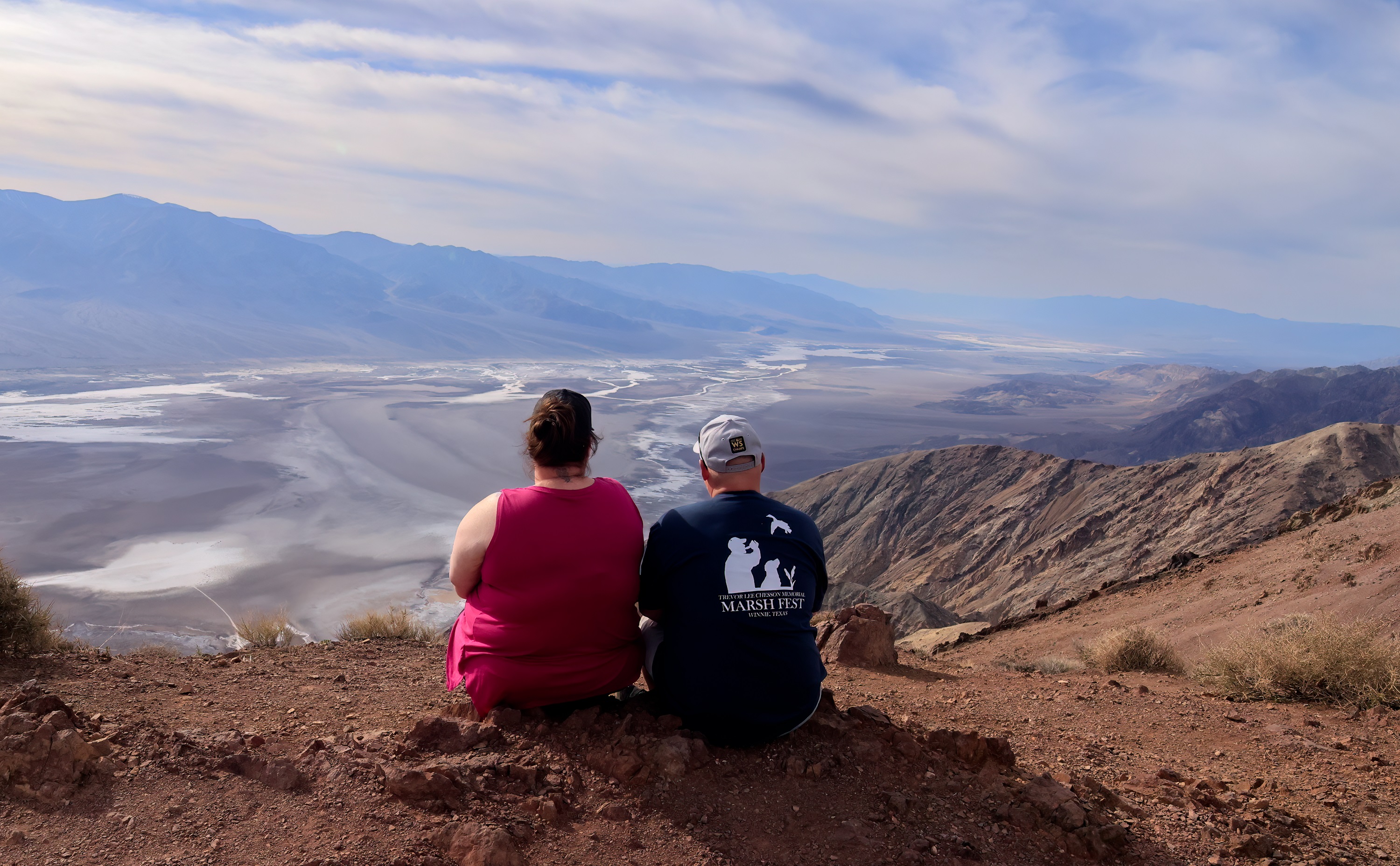 Two people sit on the ground with their back to the camera. A broad white and brown valley is beyond them.