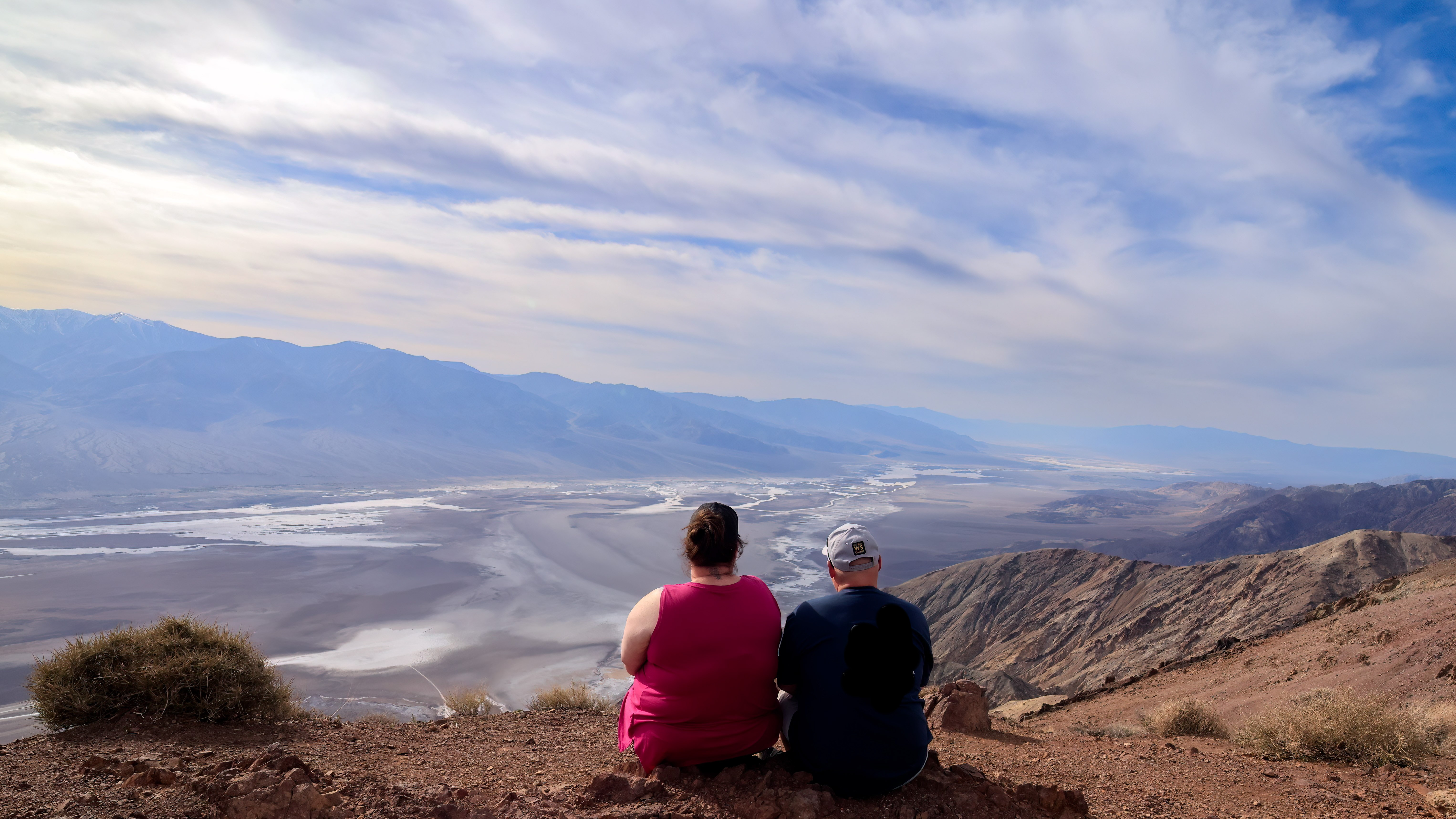 A person a pink shirt and a person in a black shirt sit looking over a wide, flat, brown and white valley.