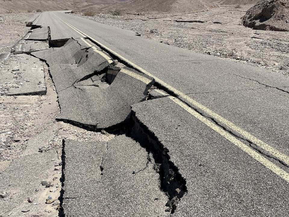 An asphalt road is a few feet above the surrounding gravel wash. The left lane's asphalt pavement is broken into large pieces and collapsed.