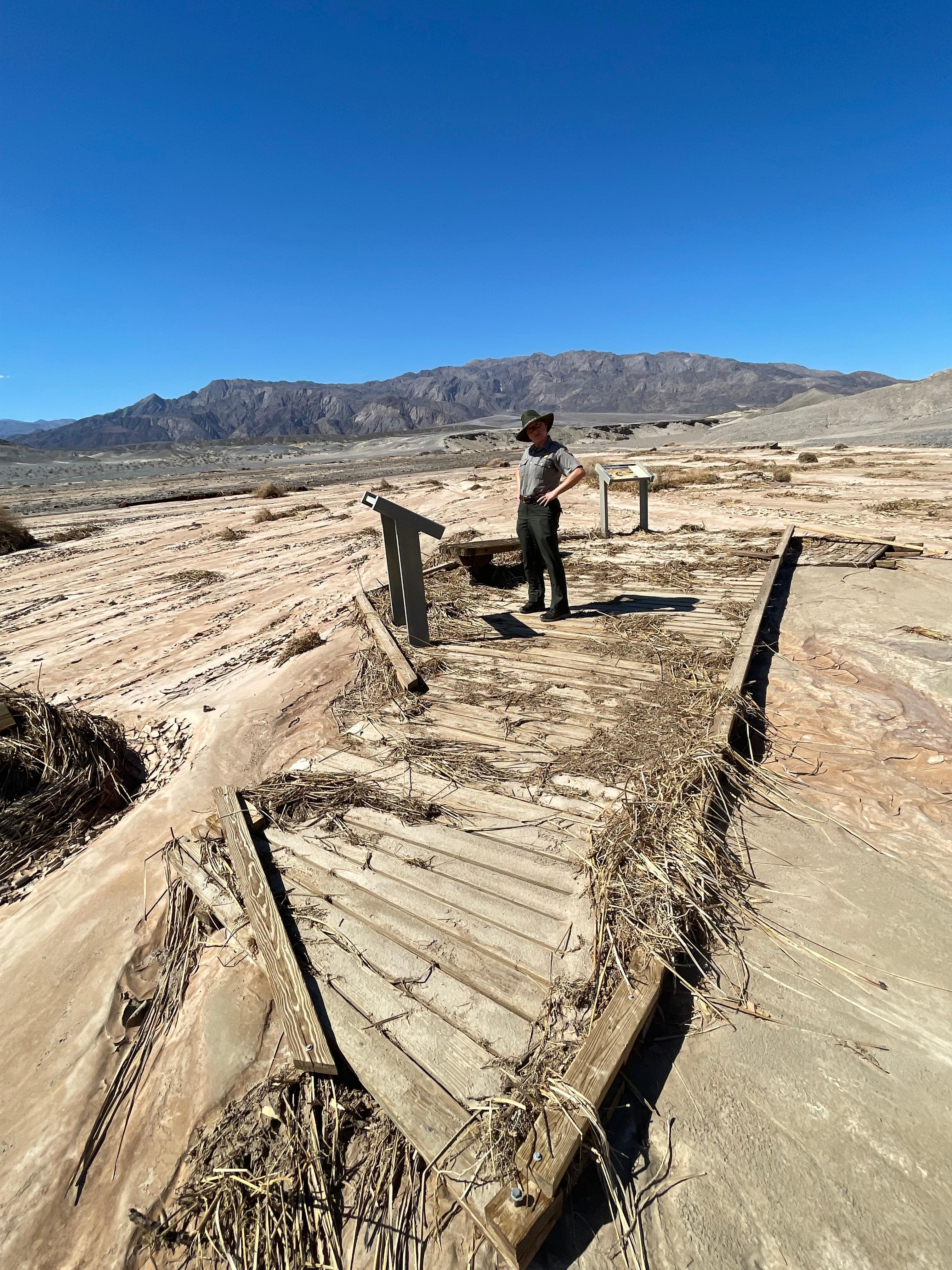 A park ranger wearing dark green pants and gray shirt stands on top of an isolated section of wooden boardwalk surrounded by a dry, barren wash.