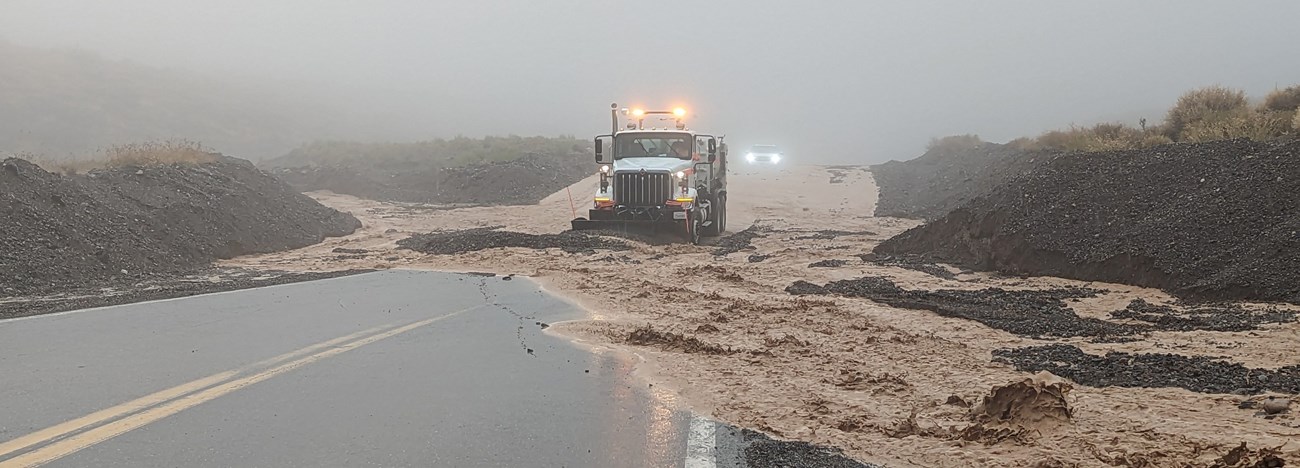 A truck pushes flood water and rocks from a road that dips between two hills on a foggy night.