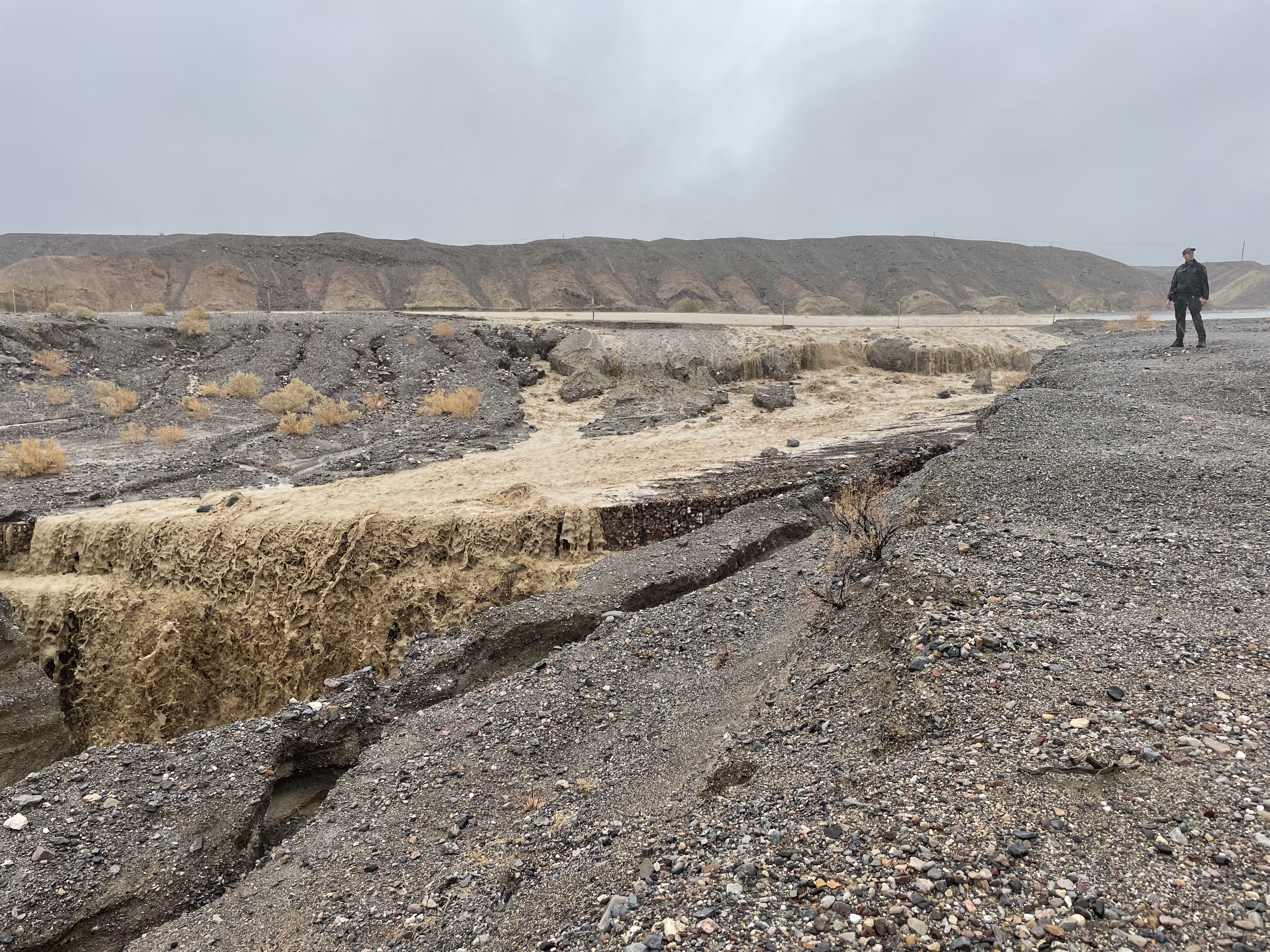 A person stands in a rocky desert landscape next to a newly formed river of muddy flood water.