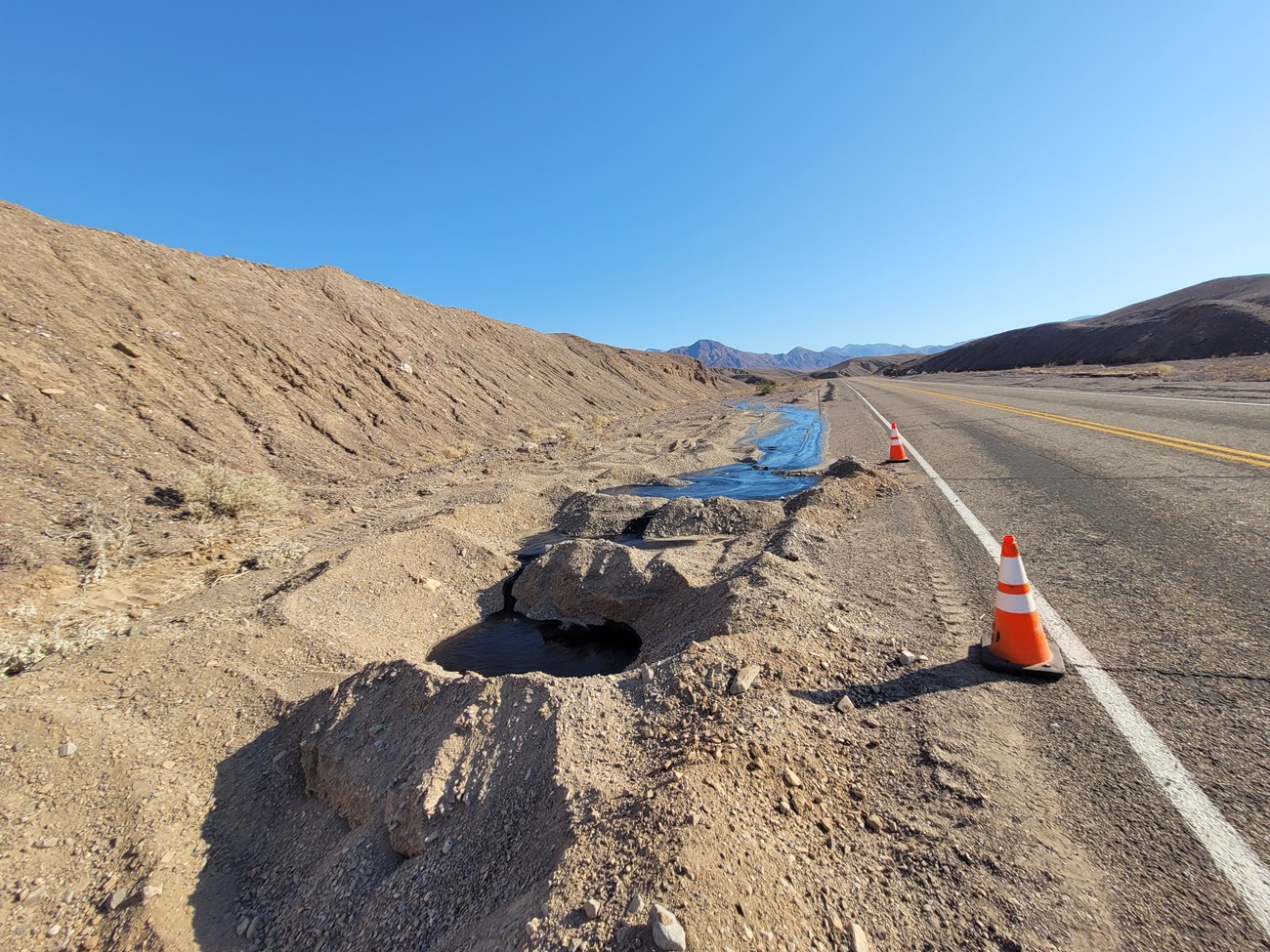 Black liquid asphalt flowing downhill toward the camera, with holes dug in foreground.