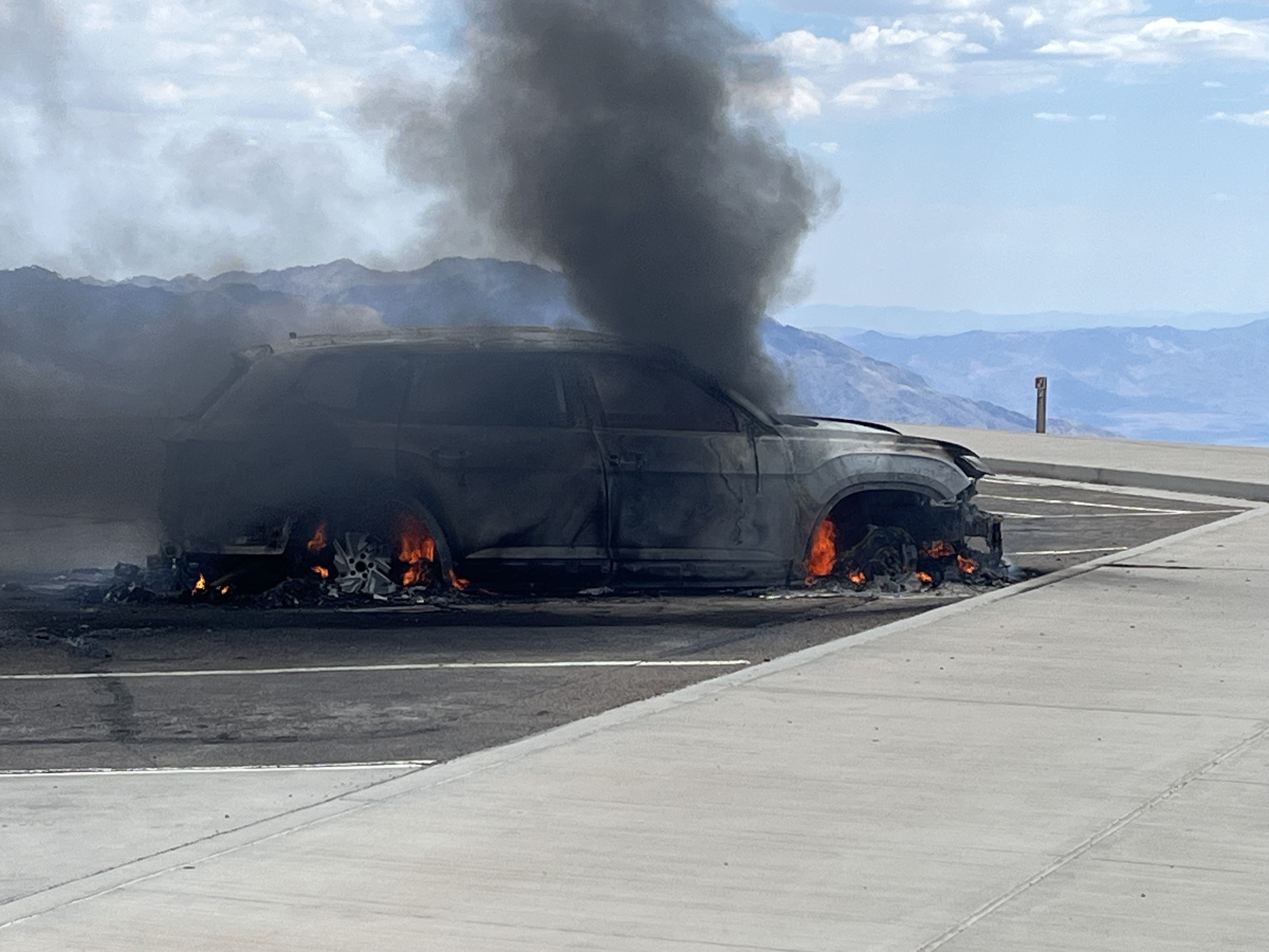 Black smoke partially obscures a gray vehicle with flames in its wheel wells. Mountains are visible in the distance beyond the end of the parking lot.
