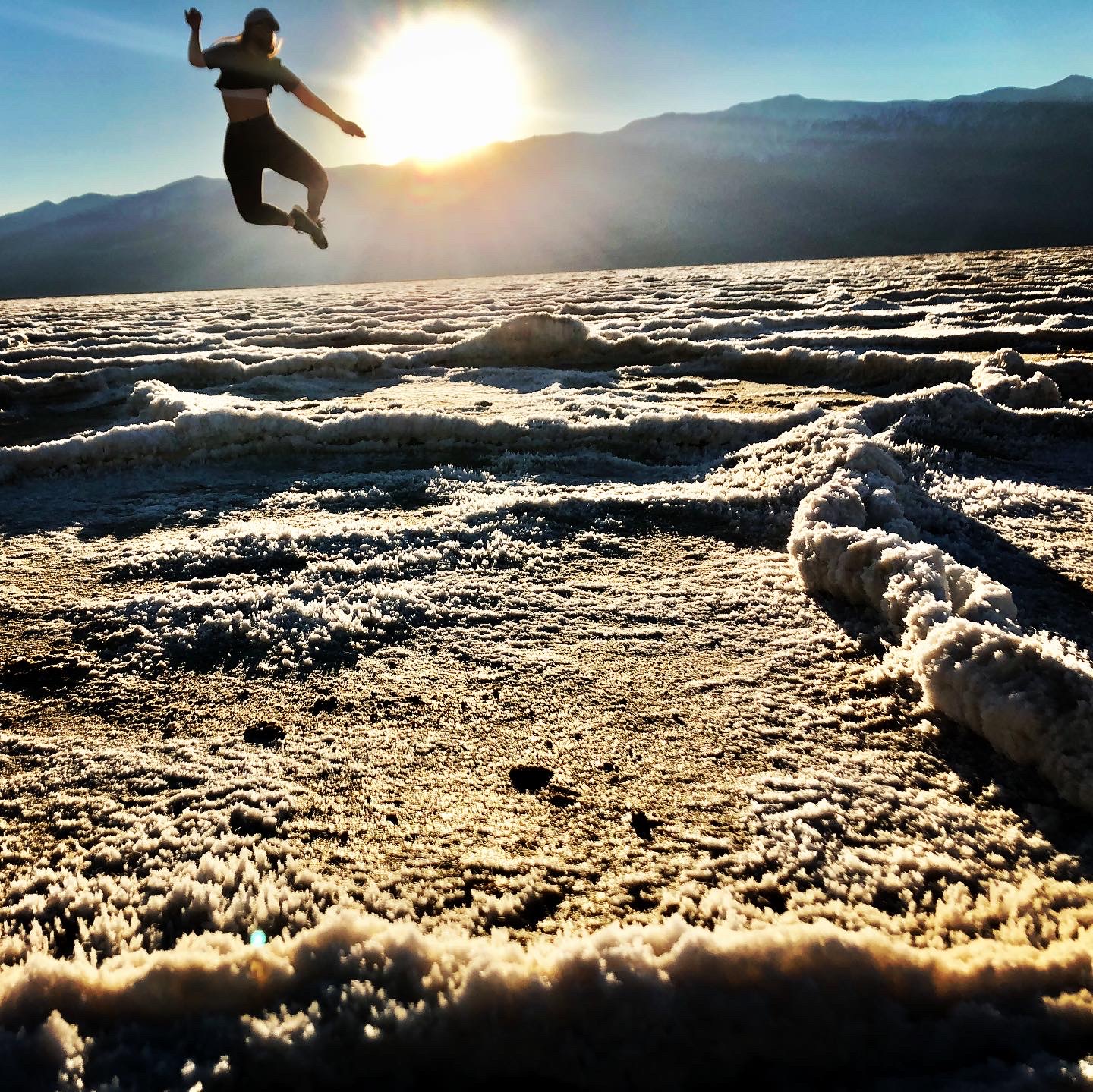 A woman leaps into the air. The foreground is a salt flat with polygon shapes, and a setting sun and mountains are in the background.