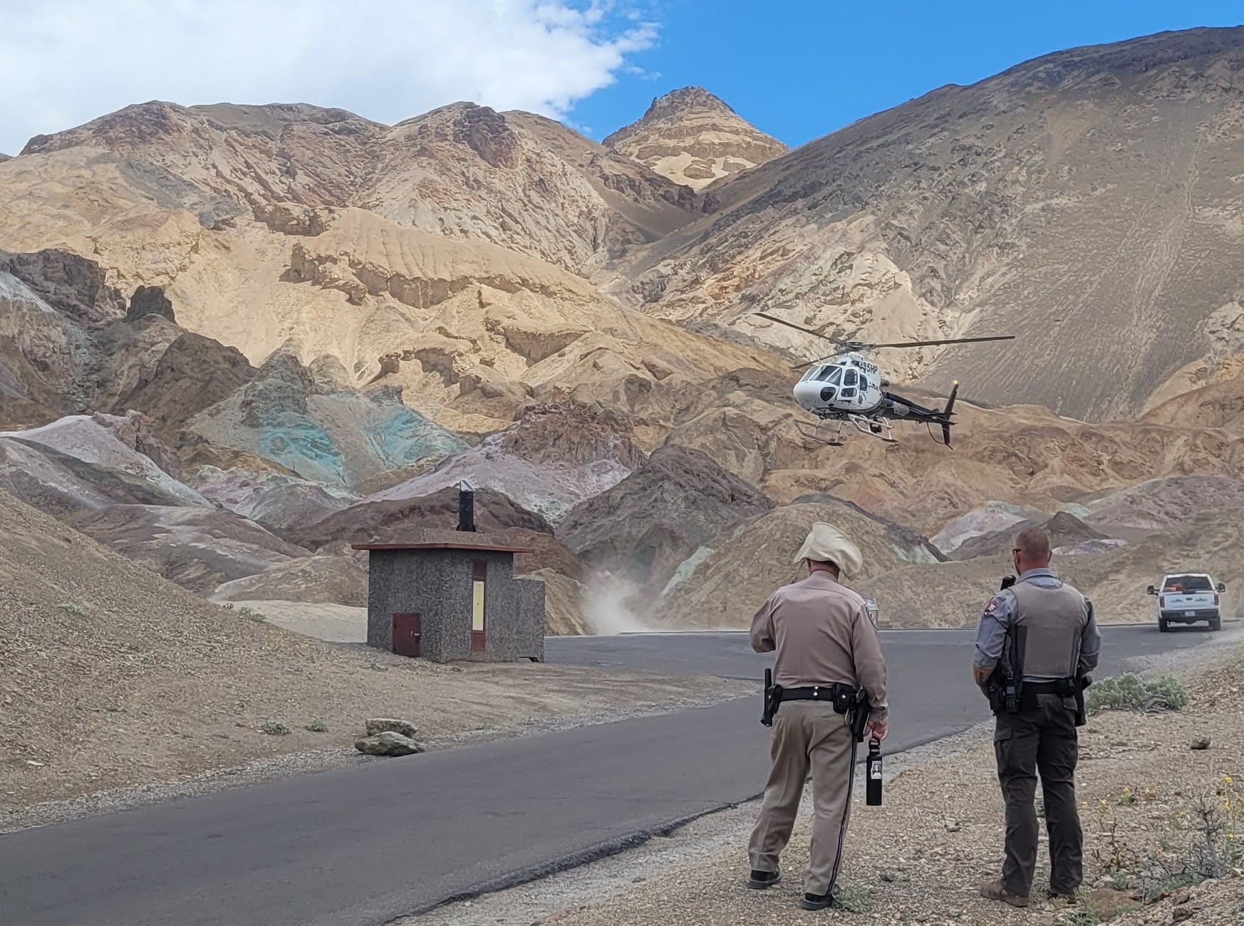 Two uniformed men stand with looking at a helicopter landing in a small parking lot next to a small building. Hills in the background are brown, yellow, pink and green in color.