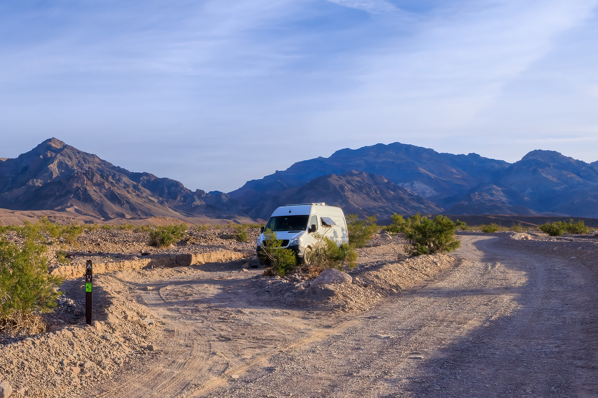 White camper van parked behind creosote bushes in a pull-through campsite along a dirt road in a desert setting with mountains in the background.
