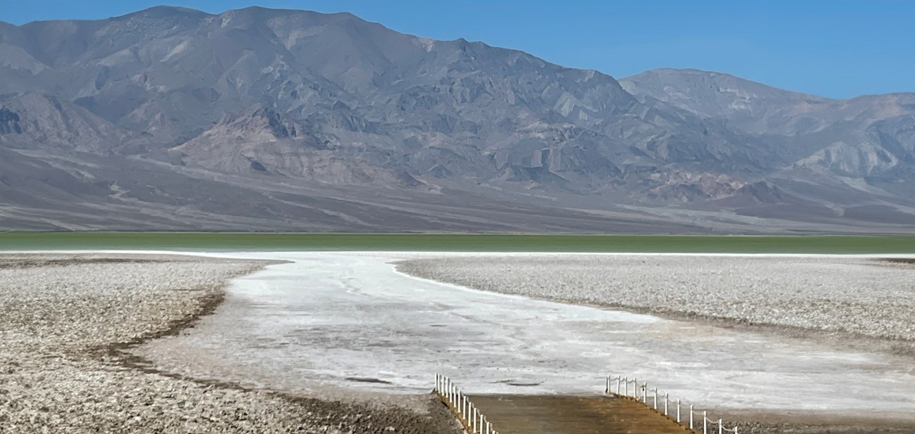 A wooden boardwalk stops at the edge of a salt flat that has a shallow green lake on it with mountain landscape in the background.