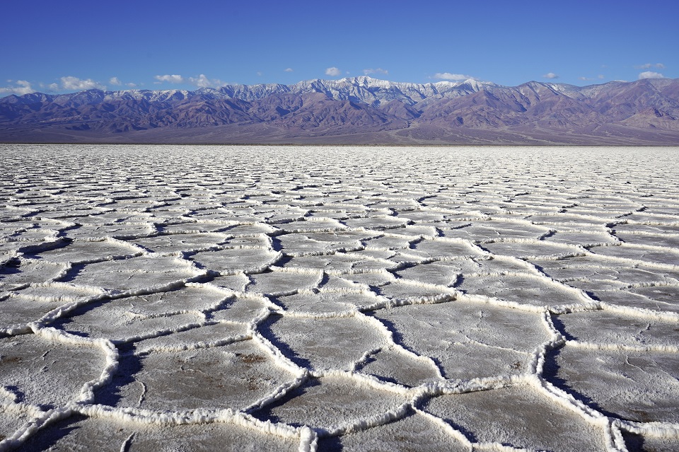 White salt ridges surround flat polygons in the foreground. Brown mountains with snow on top are seen in the background below a blue sky.