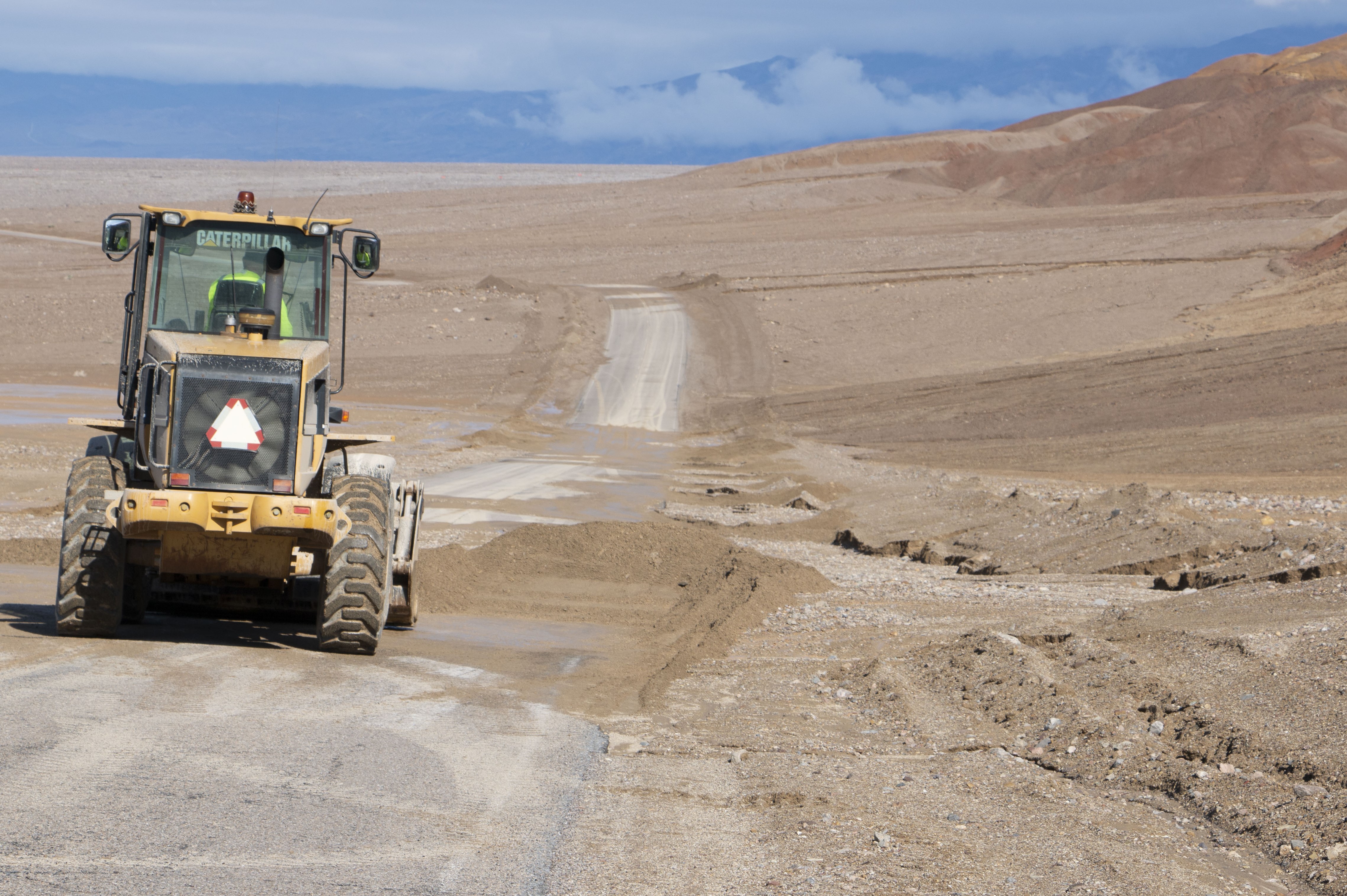 A yellow and black front-end loader is seen from the back as it scoops mud off a desert road.