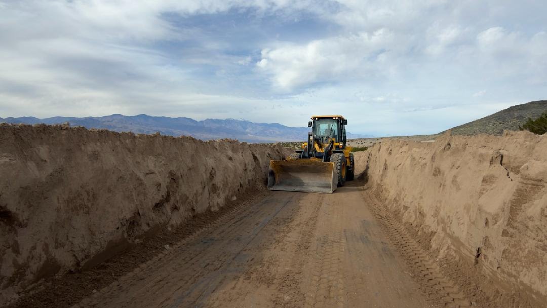 A yellow front-end loader sits in a narrow trench of between walls of sand.