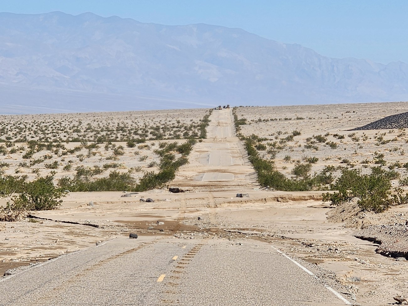Photo of a road partially covered by sand and gravel. Just the top of a white speed limit sign is sticking out of the debris on the right side of the road.