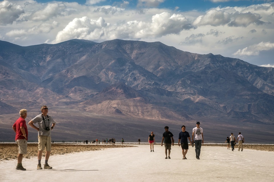 Visitors stroll from Badwater out onto the salt flats. They are walking on a broad, white path of compacted salt. Tall mountains are seen in the background.