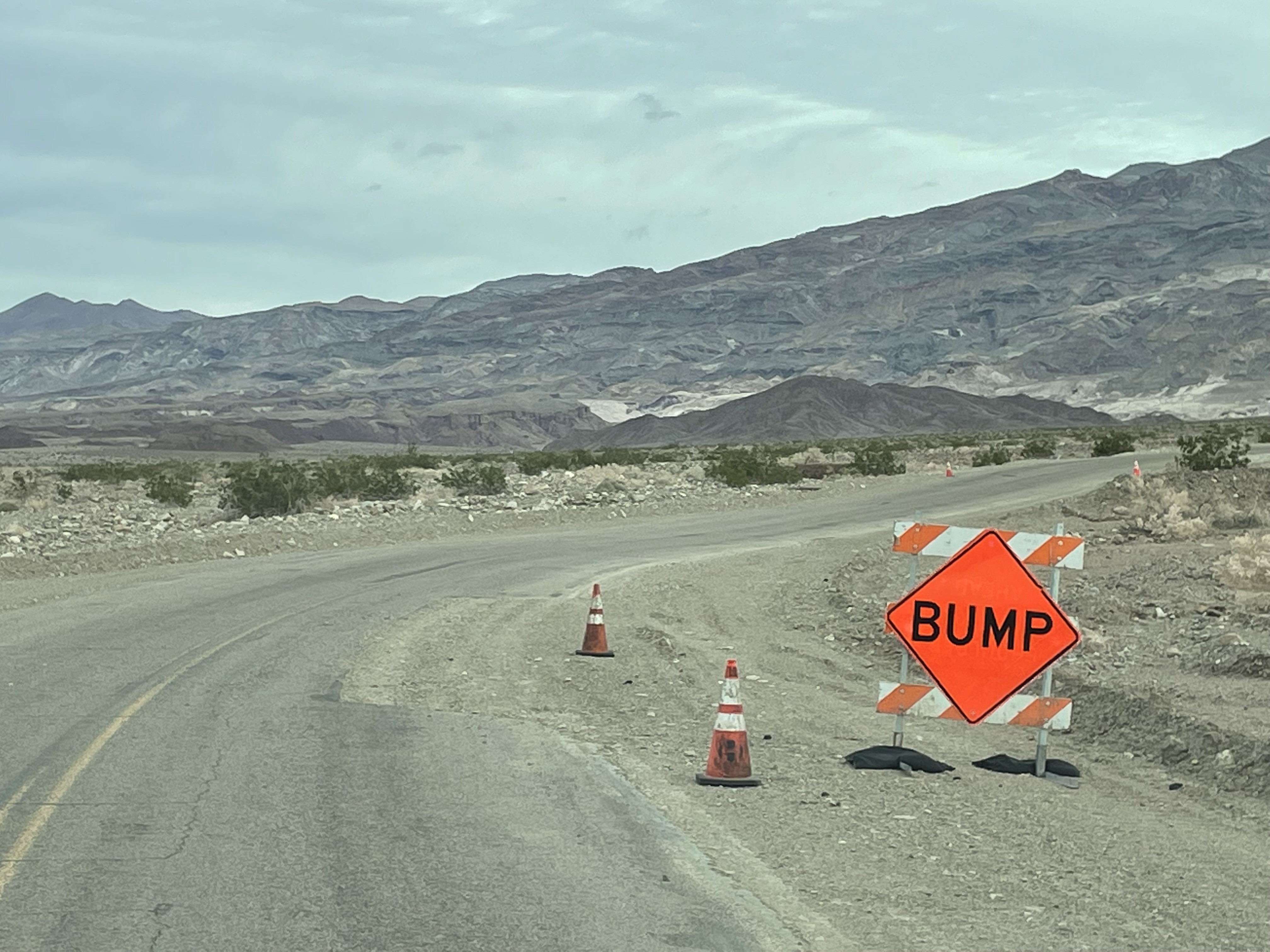 A paved road curves from bottom left to the right of the photo. An orange sign says "BUMP". Just past the sign is a section where half of the pavement is missing.