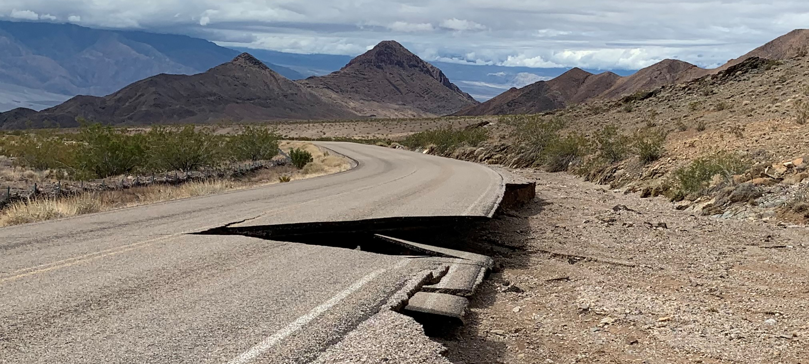 Damaged asphalt on a road that winds through a hilly landscape leading up to a mountain landscape.