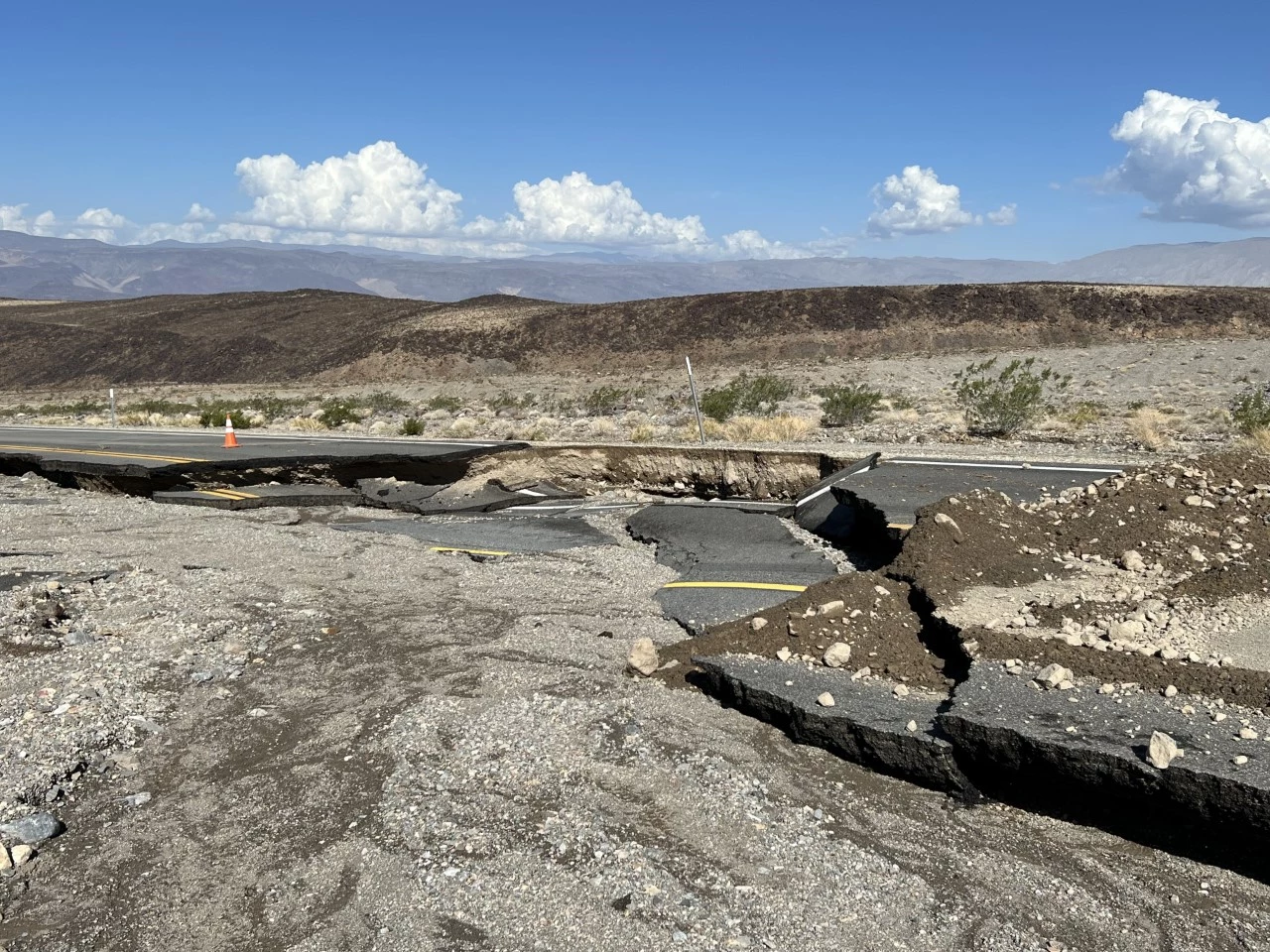 A damaged paved road in the foreground shows two-foot-deep missing sections of pavement across the entire road.