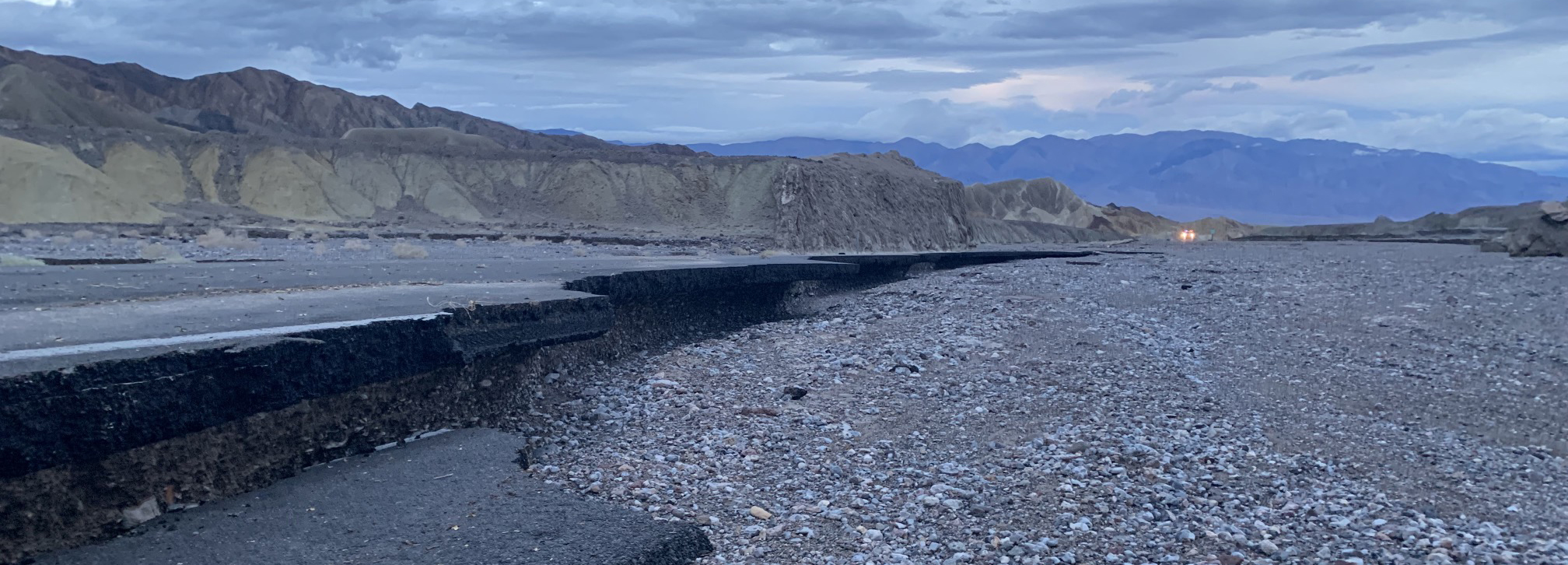 Badlands and mountains line a road undercut by rocky debris caused by floodwaters.