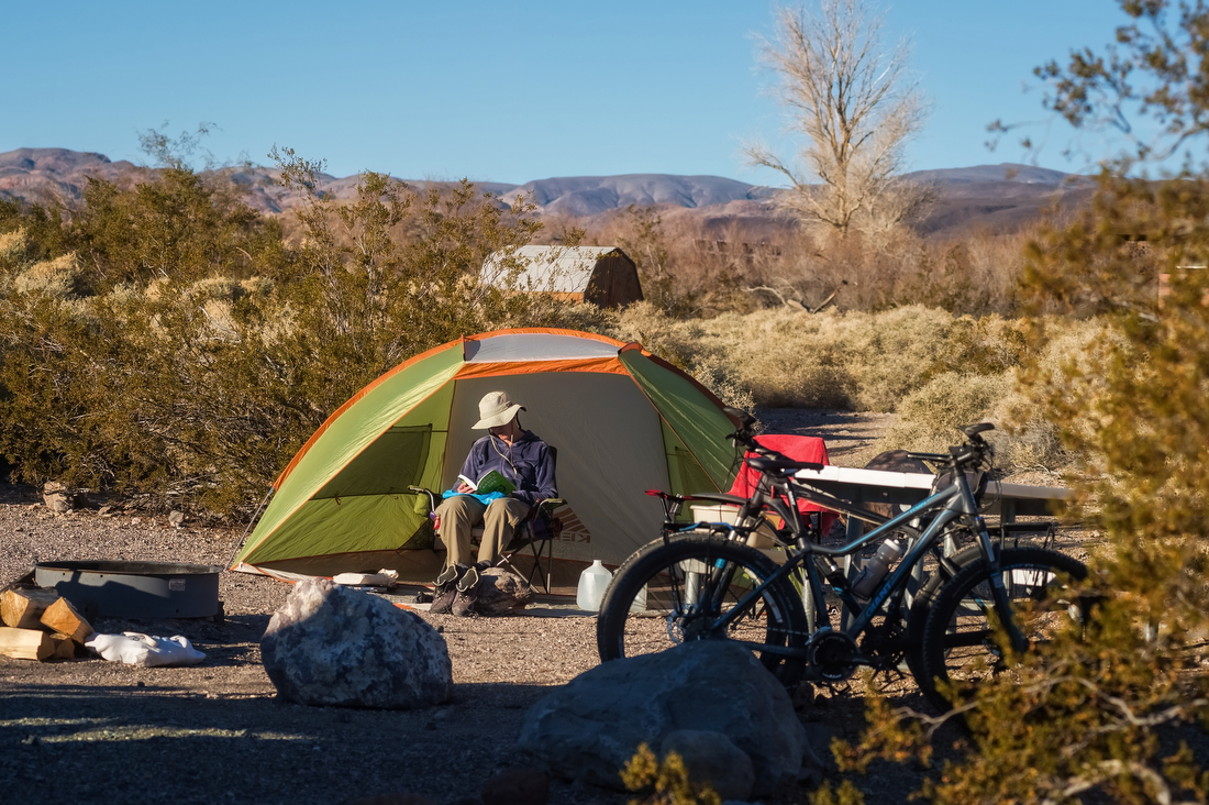 A camper sits in a chair in front of a tent with camping and recreation gear laid about.