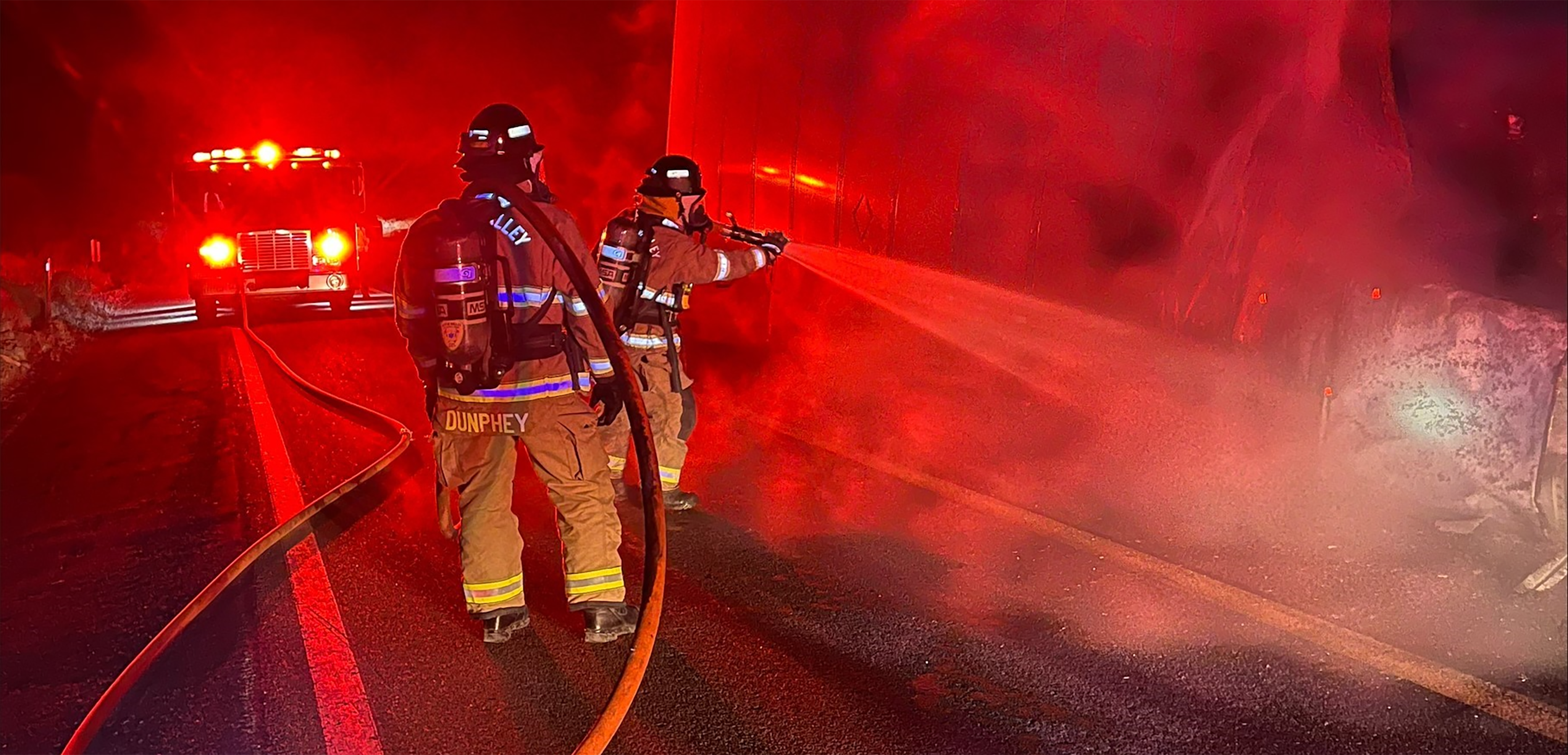 Image of uniformed firefighters putting out a semi truck on fire at night, the image is lit with red lights.