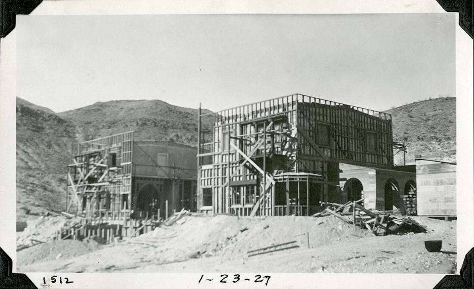 Black and white photograph of a two-story rectangular building under construction. It is surrounded by scaffolding and is missing the middle third of the building.