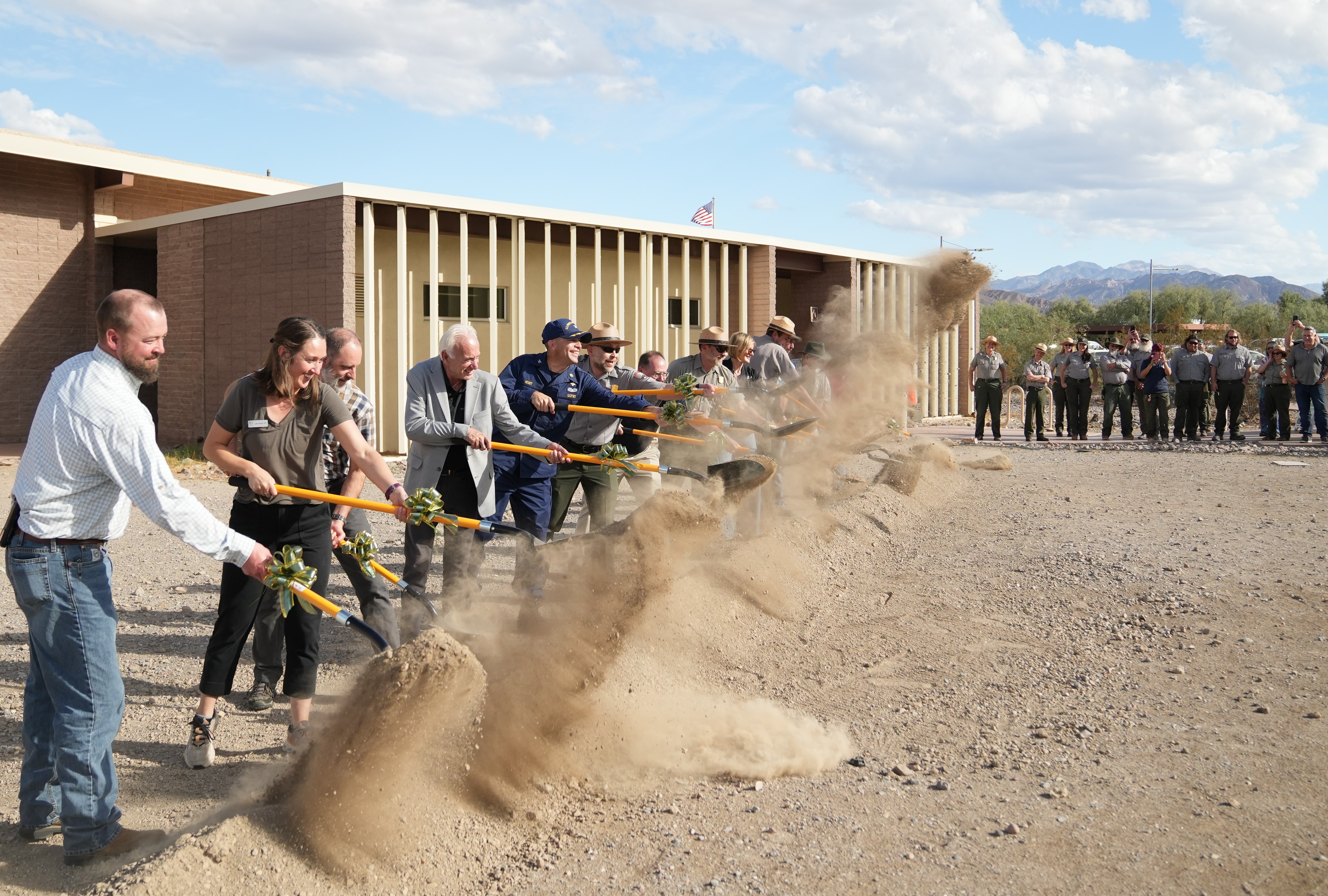 A line of people holding shovels with yellow handles throw a cloud of brown dirt while people watch.