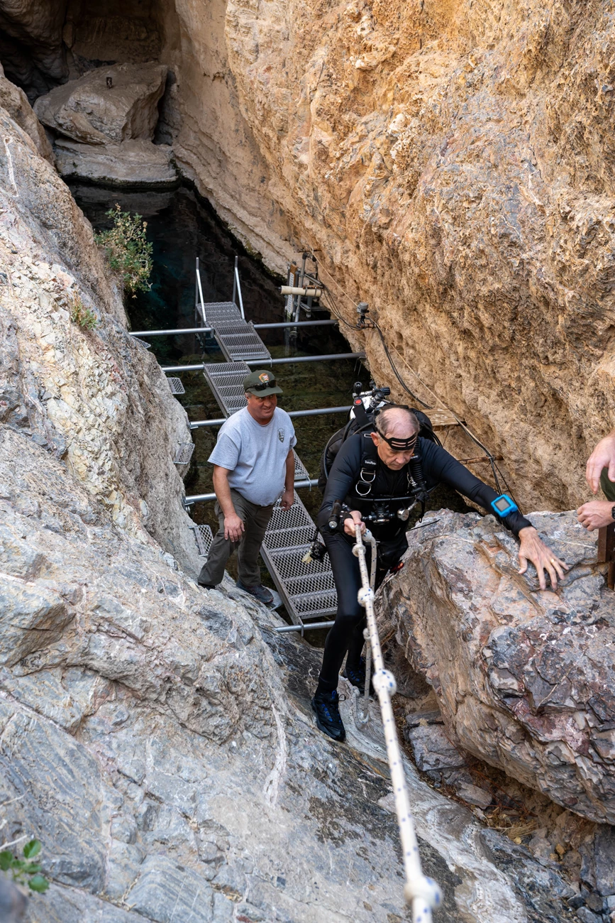 A diver climbs a rocky slope with assistance from a rope, while another individual stands nearby on a metallic platform.