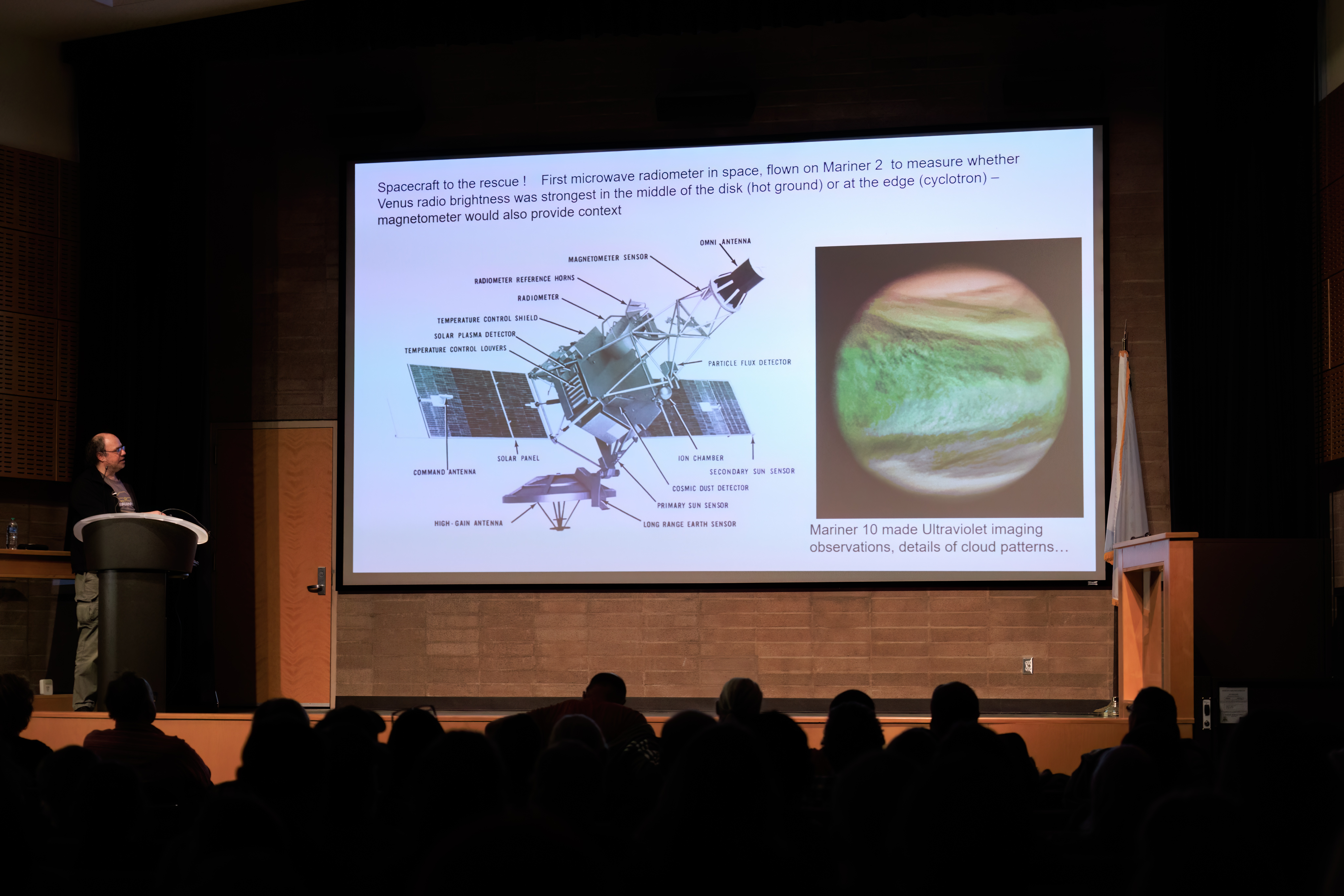 A man stands at a podium on the left side of the stage. A slide with a white background includes a diagram of a Mariner space probe, words, and a red and green ultraviolet image of Venus.