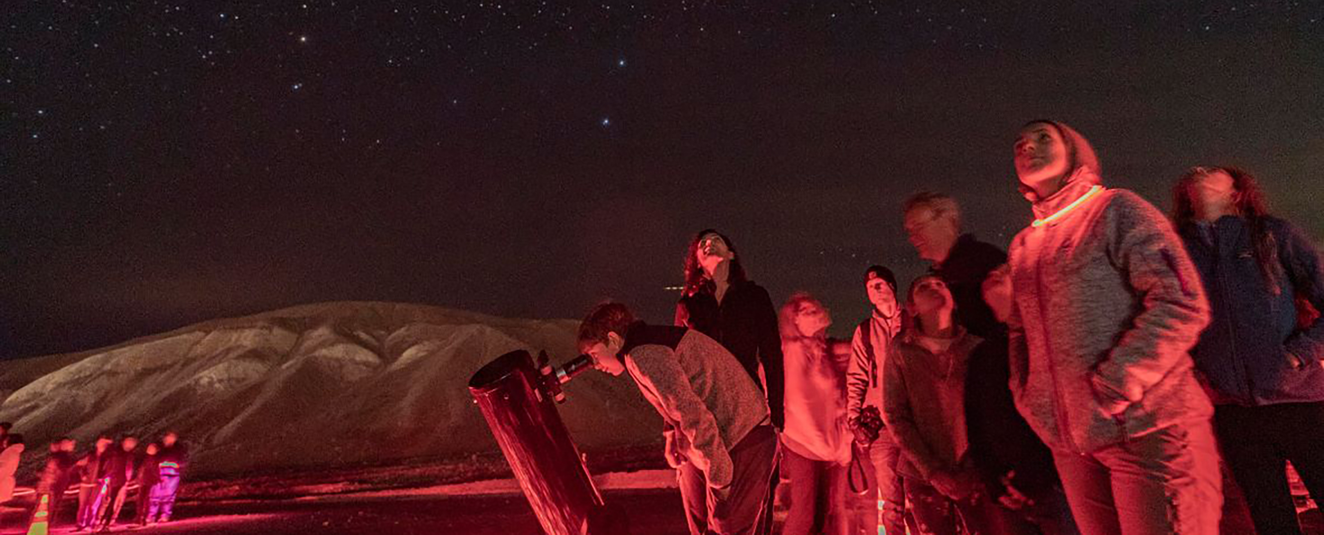 Visitors looking through a Newtonian telescope and staring at a sky full of stars surrounded by red light.