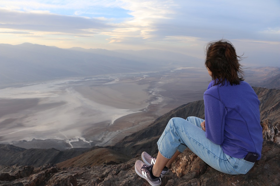 A person in a purple shirt and jeans sits with their back to the camera looking at the flat valley and distant mountains.