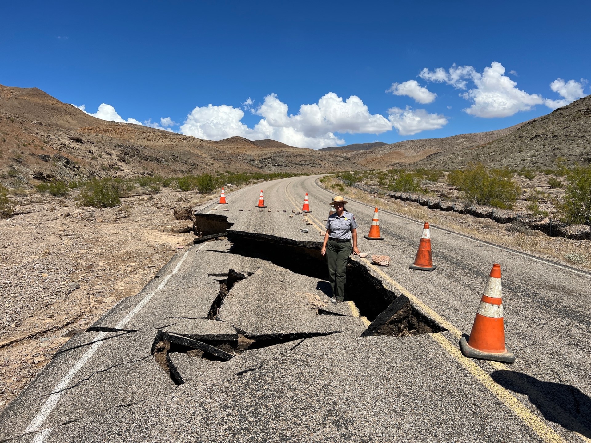 A woman in a green and gray National Park Service uniform stands on a damaged section of a paved road. Desert hills are in the background.