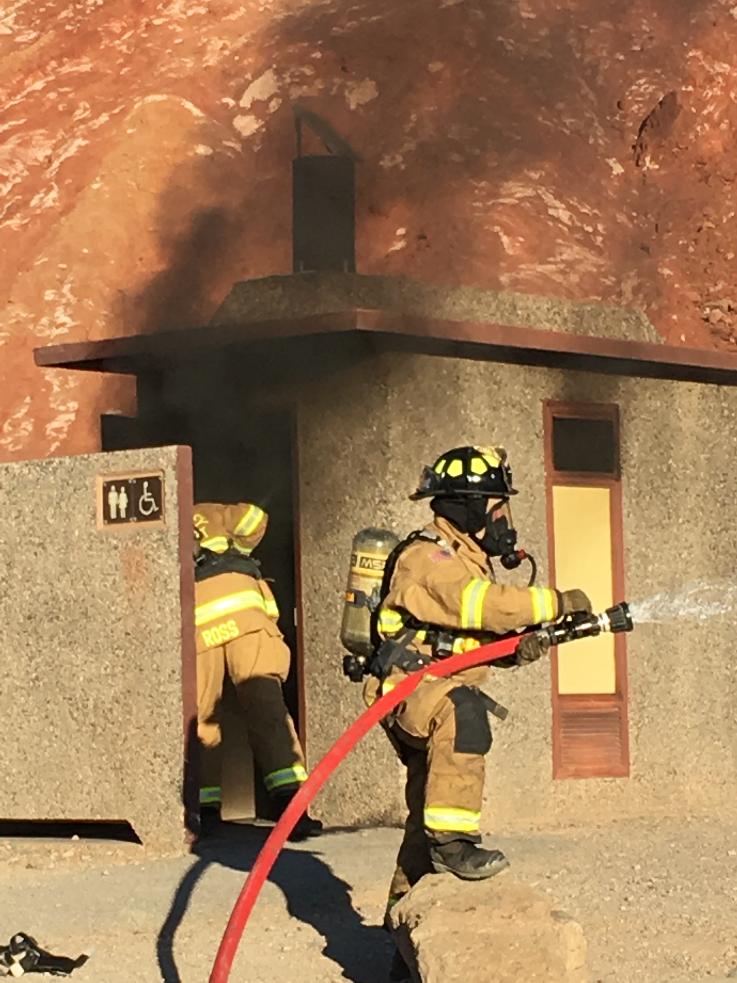 Two firefighters fight a fire coming out of a concrete vault toilet building.