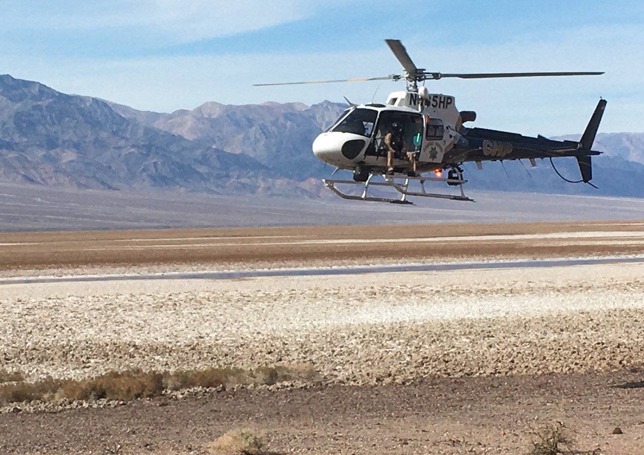 A helicopter is in the foreground in top right corner of photo. It is about 20 feet off the ground, which is gravel in foreground and salt in midground. Mountains are in background.
