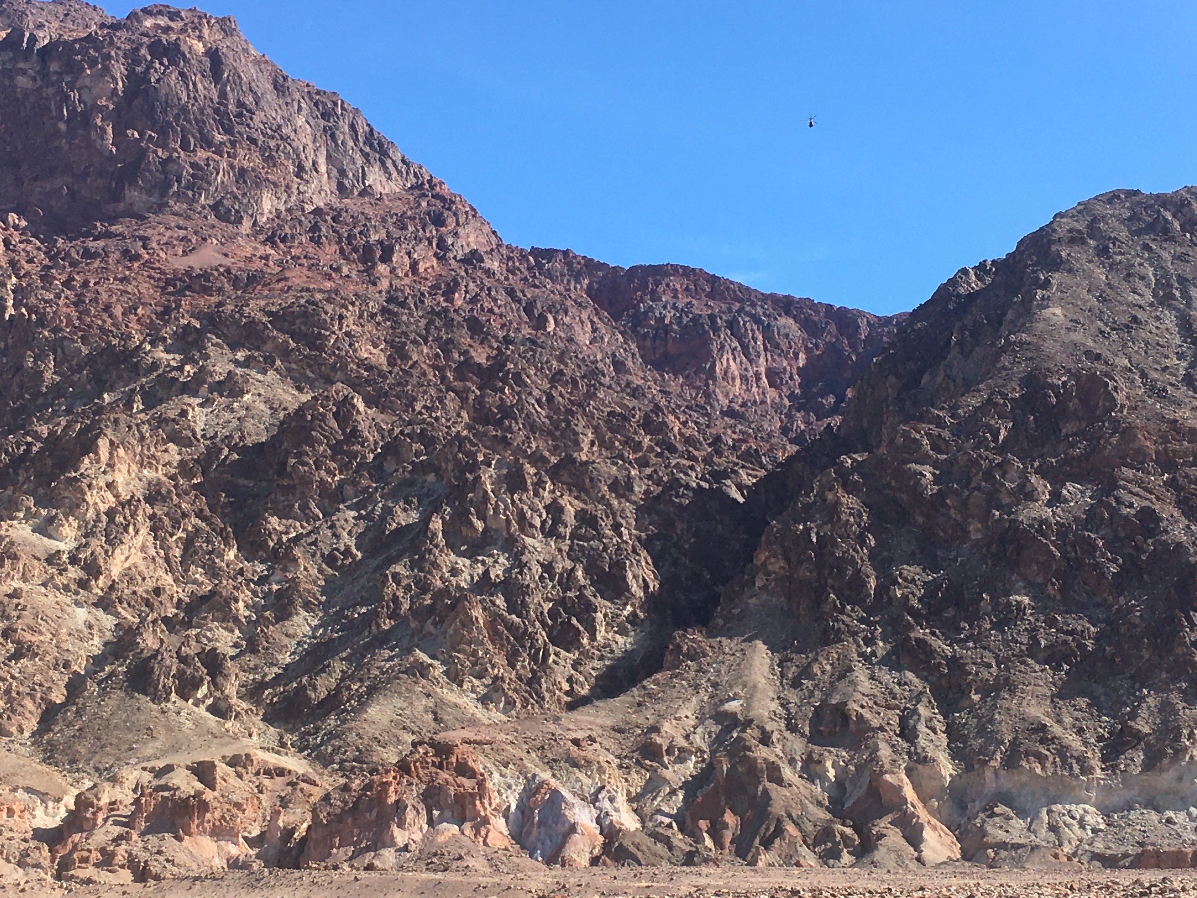 A helicopter is a small speck against blue sky over the dark brown canyon, as viewed from Badwater Road.