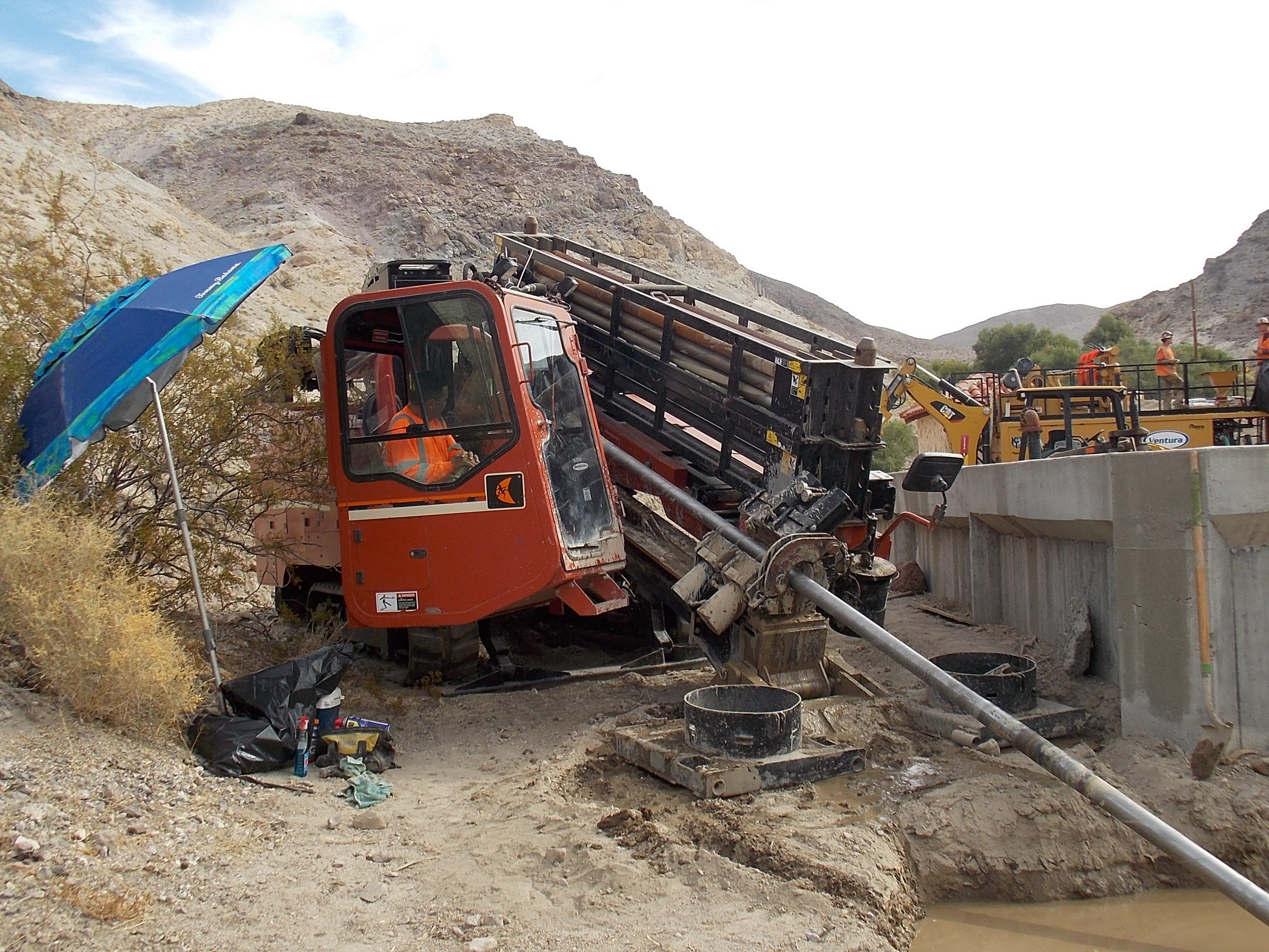 A blue umbrella is set up on the left. In the image center is an orange drill rig with a person sitting in it. The drill is going at a diagonal angle into what appears to be a mud puddle. A concrete-walled reservoir is on right in background.