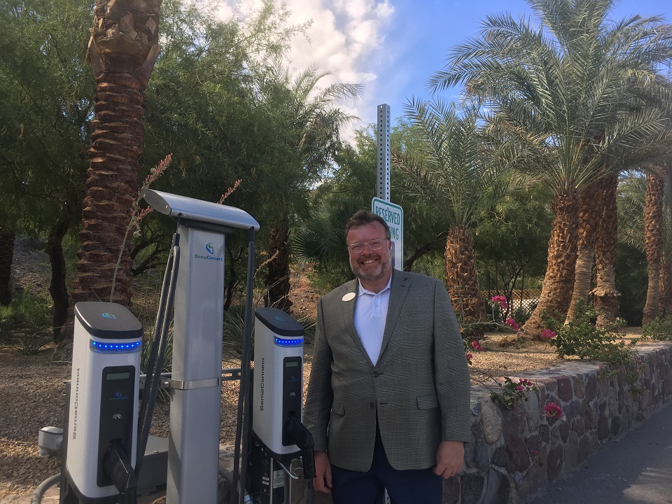Electric vehicle (EV) chargers at The Oasis at Death Valley. General Manager Trey Matheu stands next to two chargers. Behind him is a historic stone wall and lush vegetation.