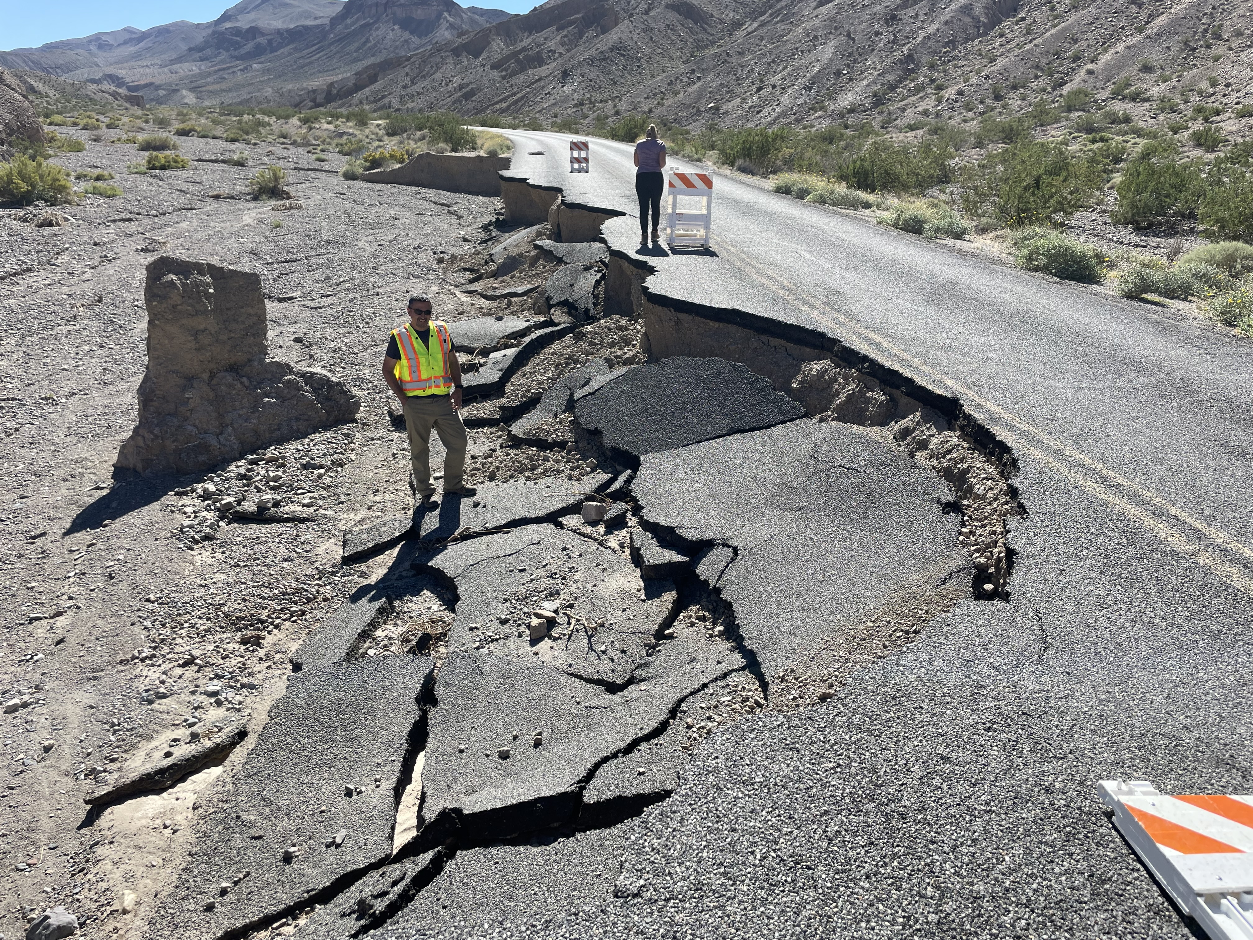 A black asphalt road heads towards mountains. The left lane is missing to the yellow center line. A person stands on the road next to a traffic sign. A second person in a bright yellow vest stands at ground level in the missing road section.
