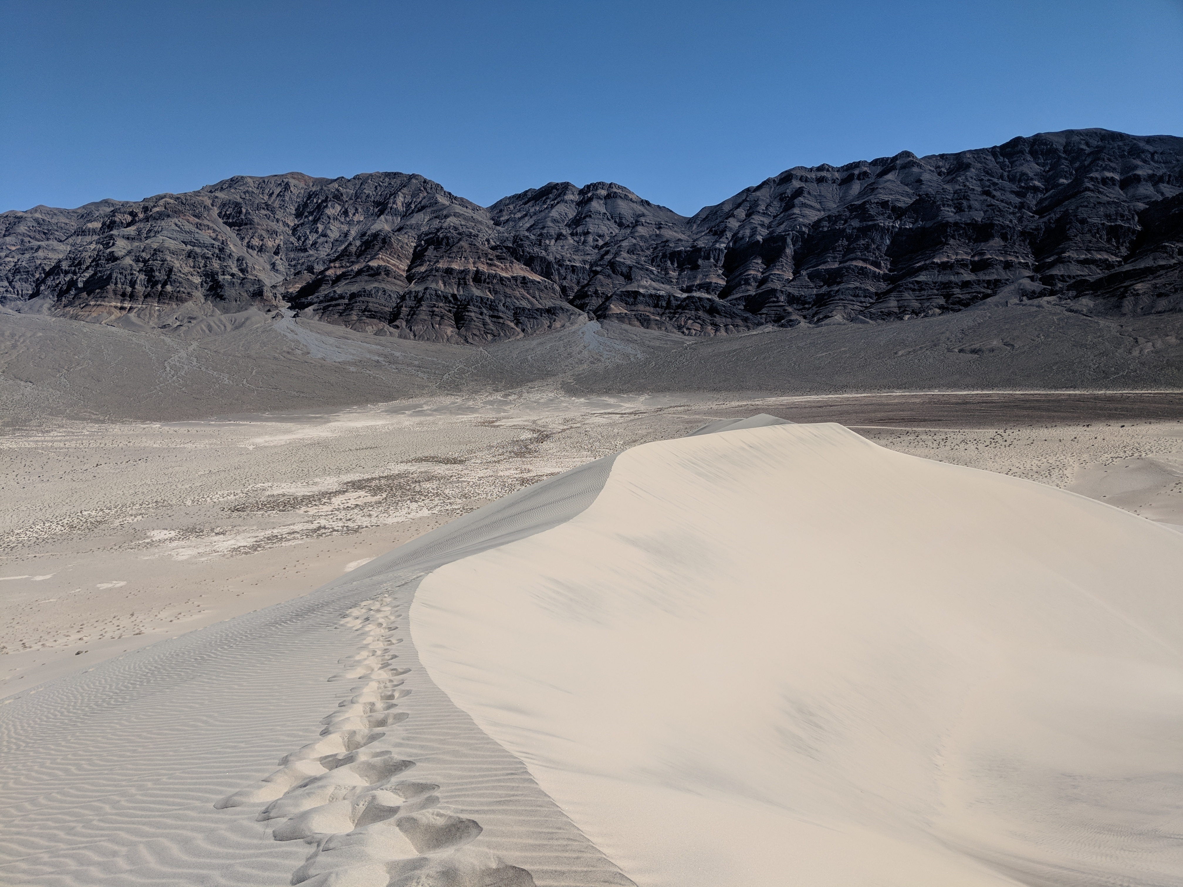 Footprints on white sand dunes in front of dark mountains.