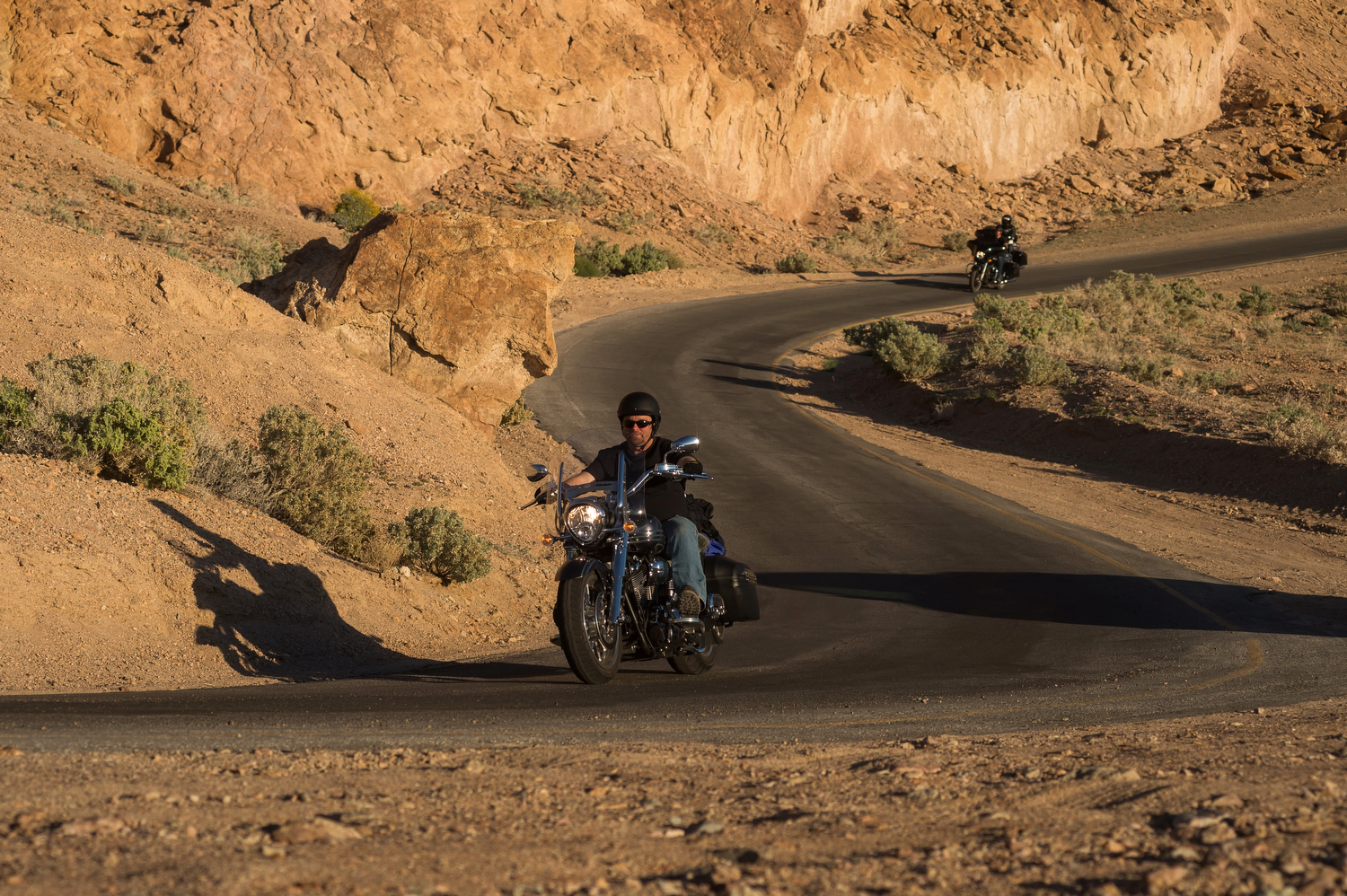 Two motorcycle drive a single-lane black curvey road through tan-color desert scenery.