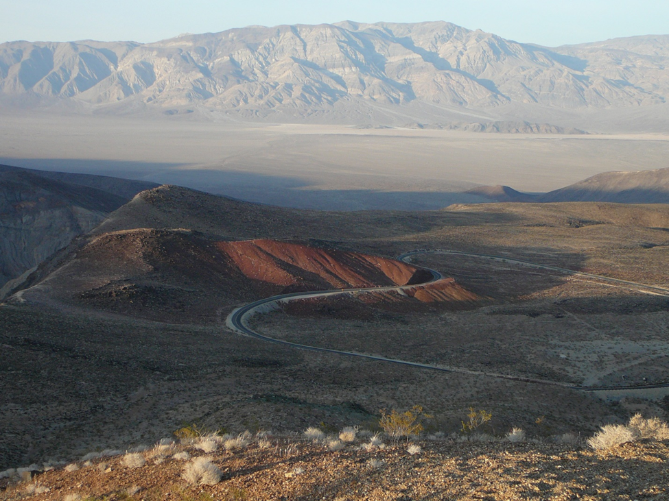View of reddish brown canyon with winding road leading into a flat area with large mountains behind.