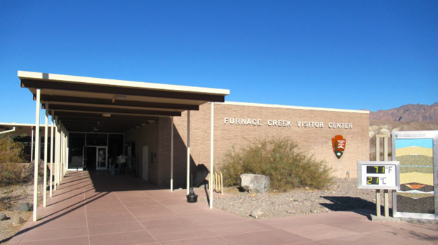 Single-story flat-roofed tan building with white awning, thermometer displaying 71 degrees F, and building name: "FURNACE CREEK VISITOR CENTER."