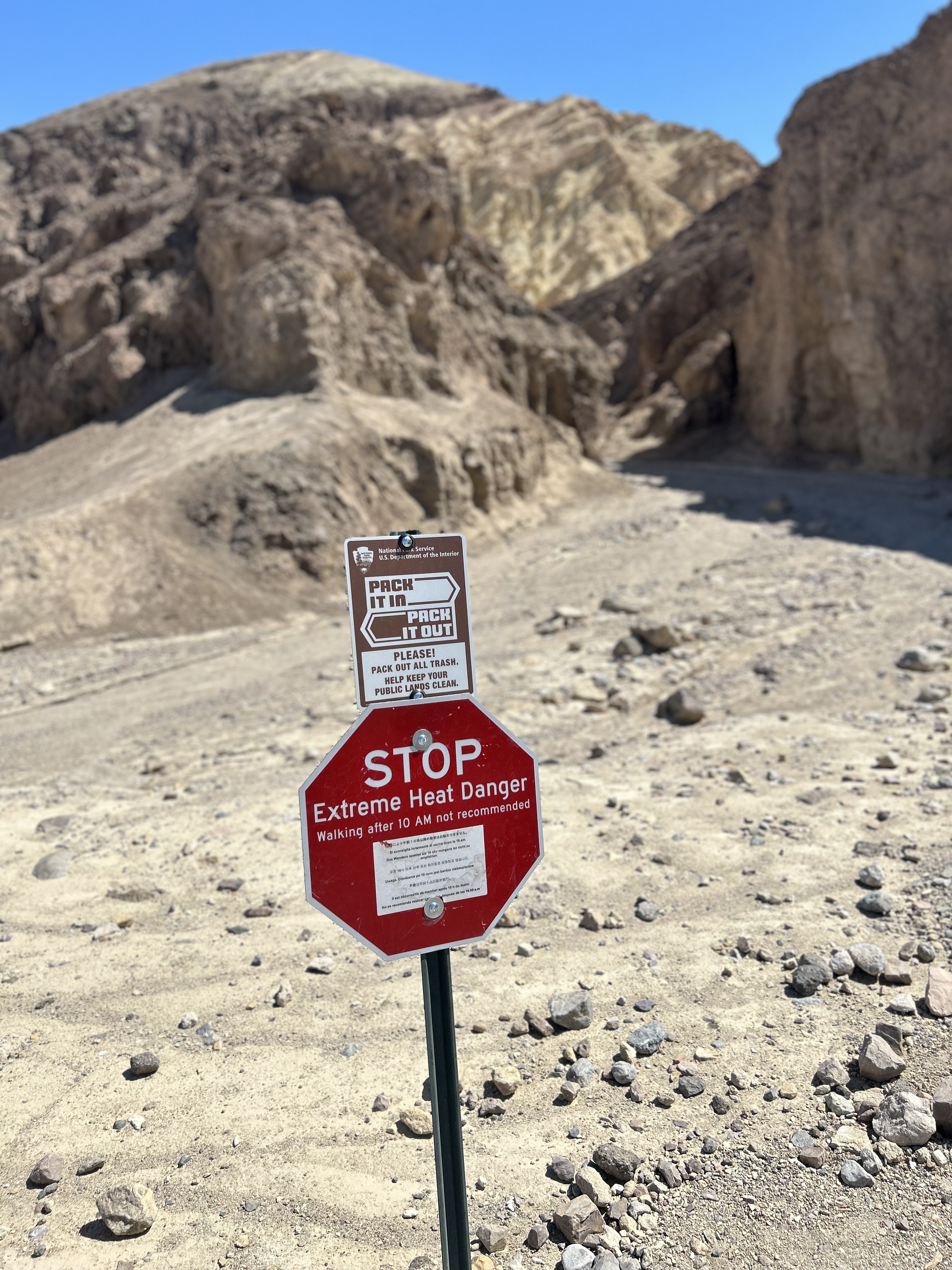 A post with signs reading "Pack it in, Pack it out" and "STOP. Extreme Heat Danger" is in front of bare rocky ground. "