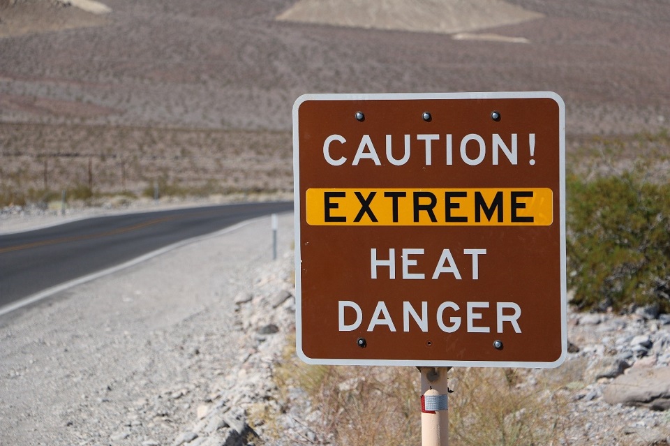 A brown sign reading "Caution! Extreme heat danger" is shown to the right of a paved road in Death Valley National Park.