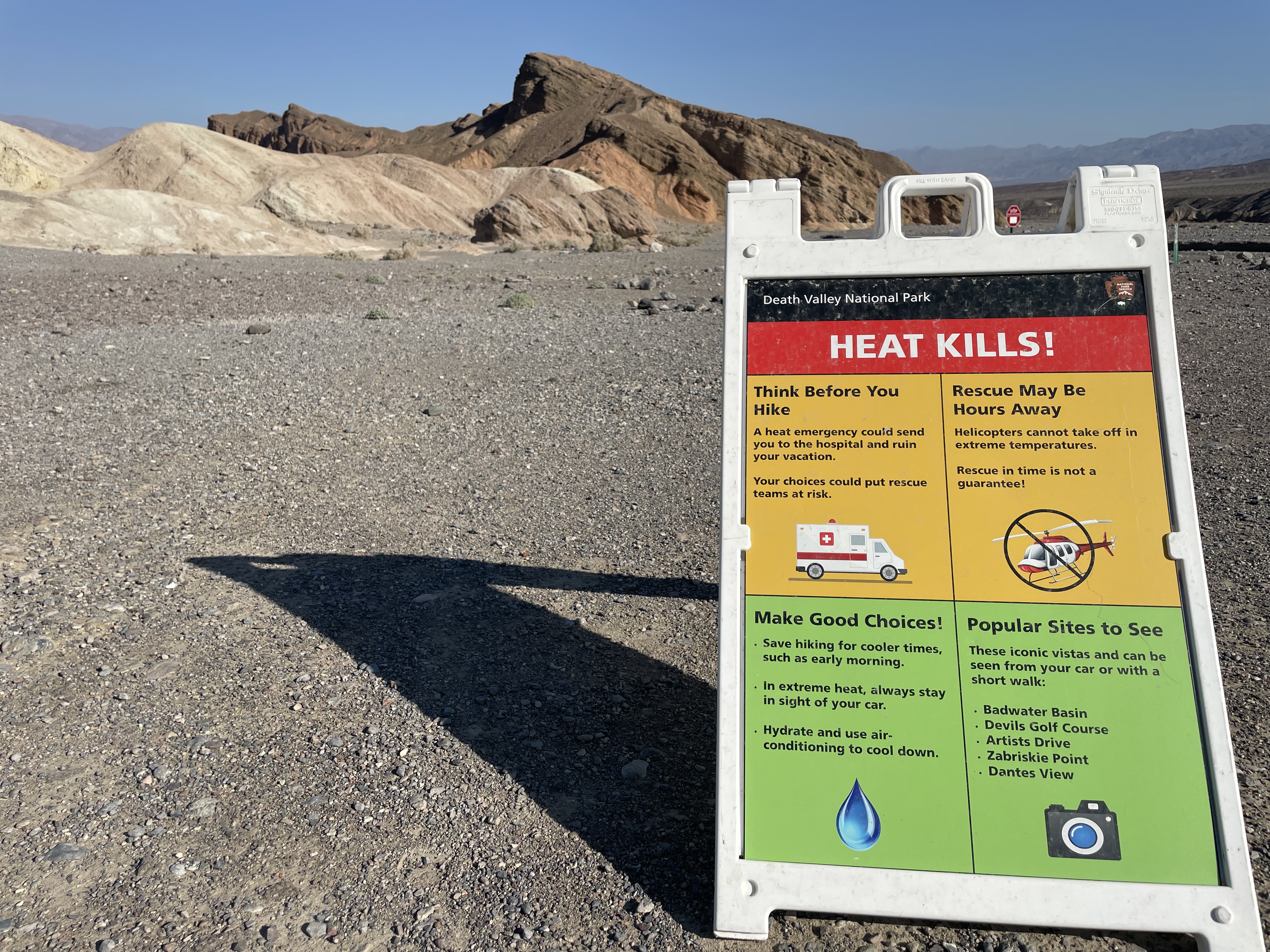 A red, yellow, and green sign in the foreground contains text about heat safety. A barren desert hill is in the background.