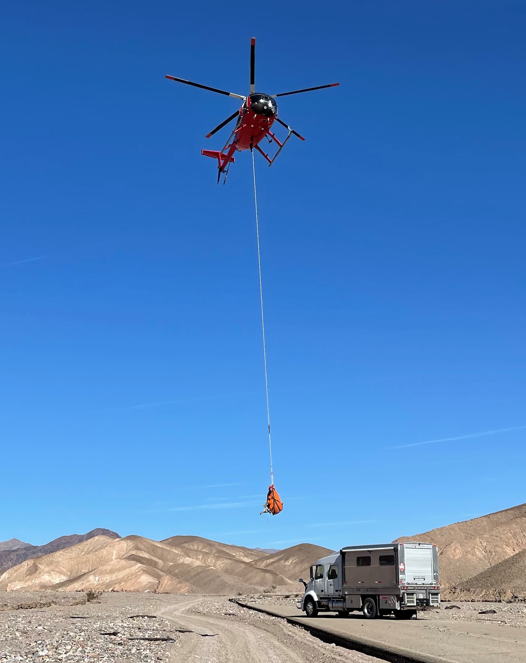 A red helicopter hovers in a blue sky with an orange object hanging by a cable from it.