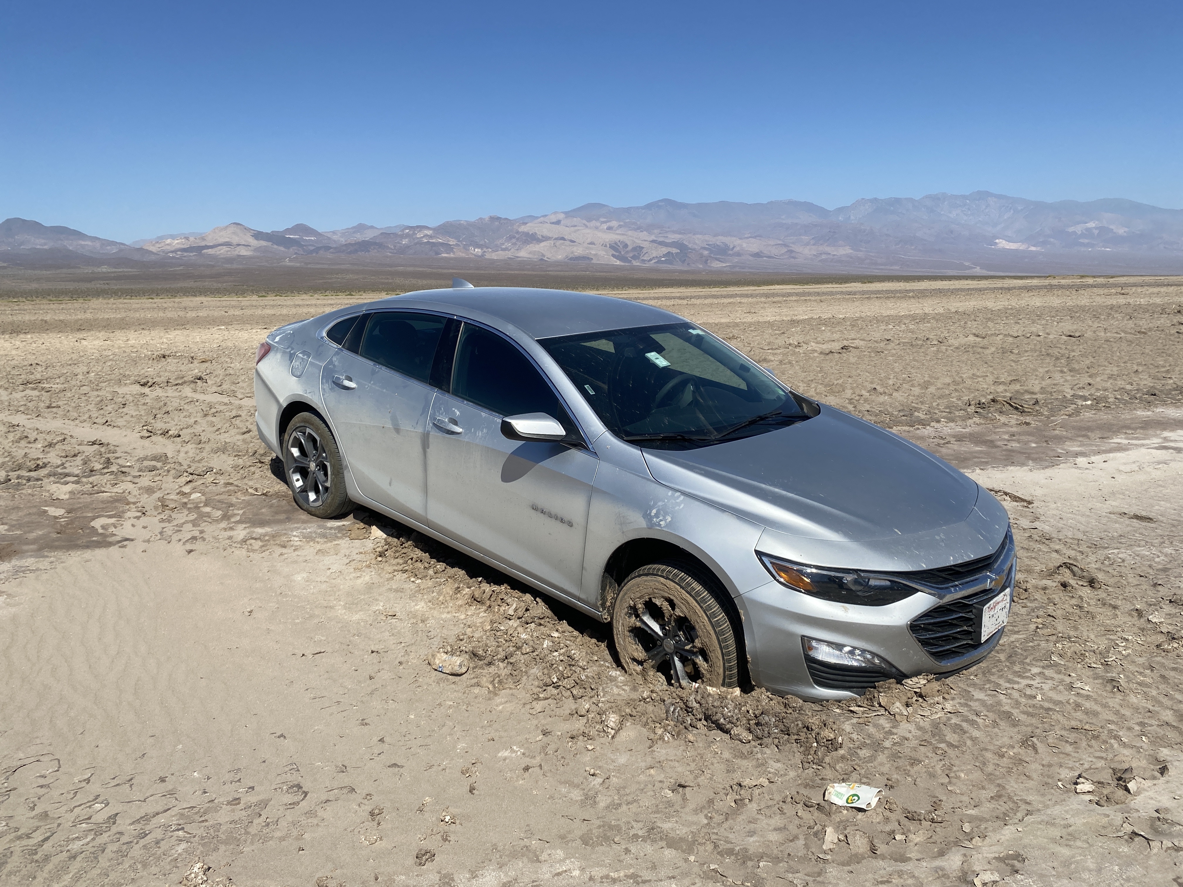 A gray car has a front tire sunk into mud. It is in a wide, flat open area with no road in sight.
