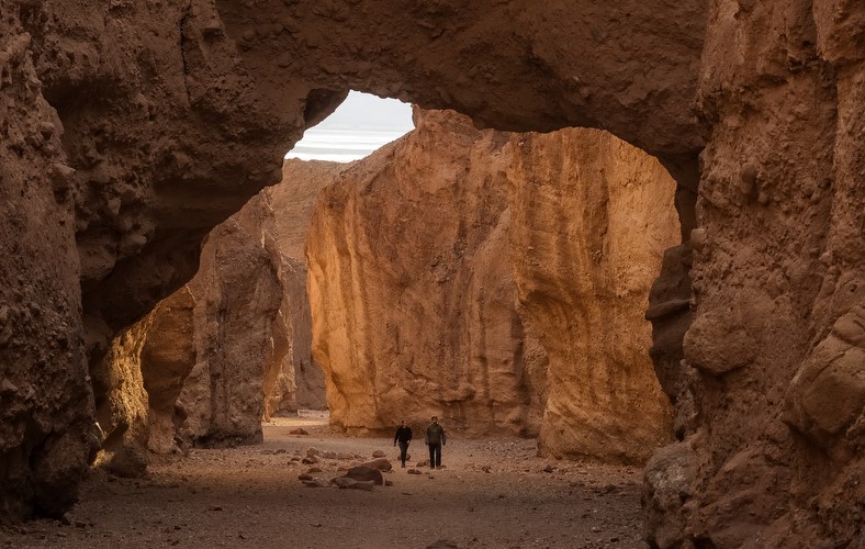 A large natural bridge frames the top of the image as two people walk toward the camera in a gray- and tan-colored canyon with vertical walls.