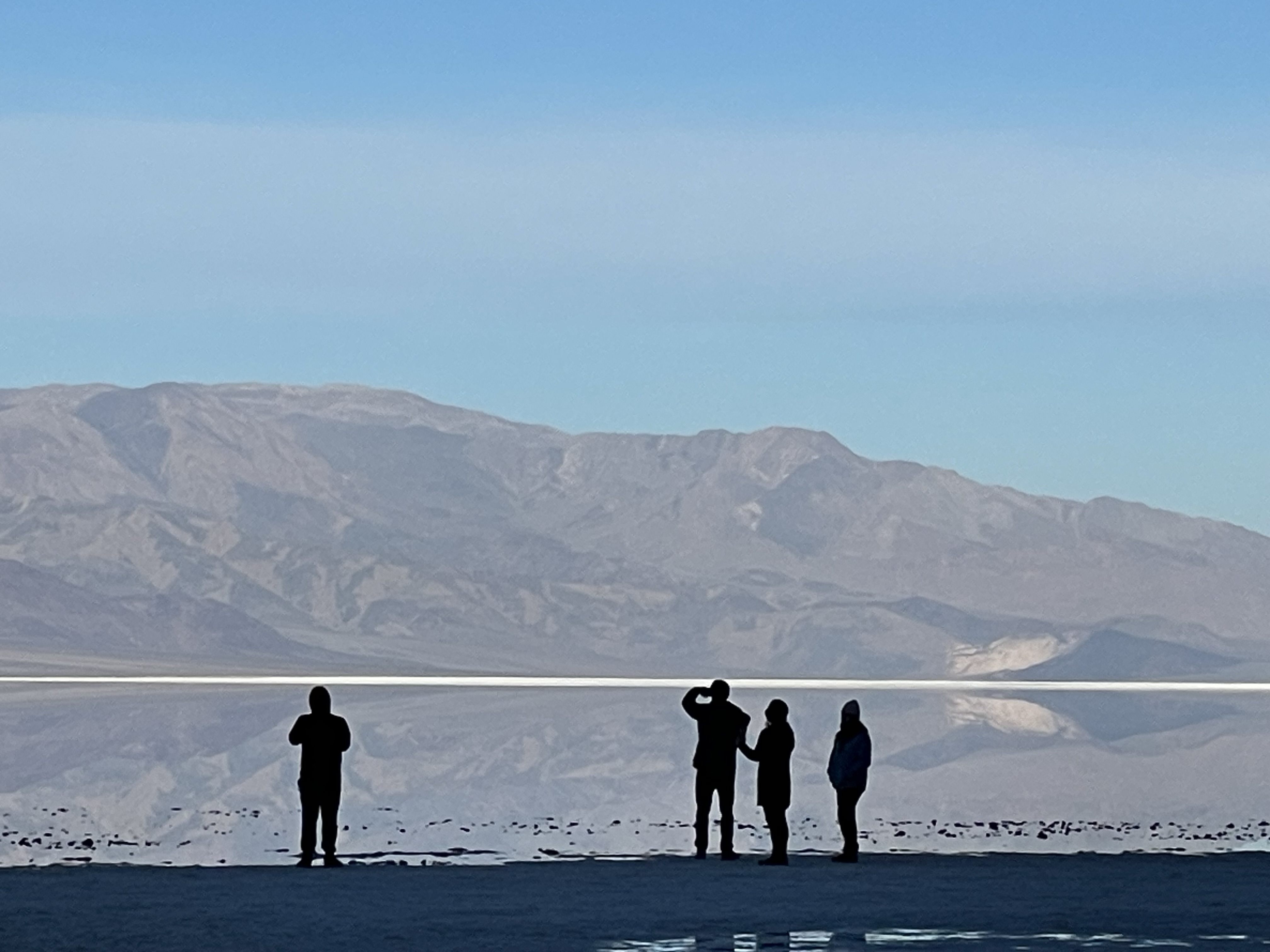 Four people are in dark silhouette looking at a brown mountain reflected in calm water.