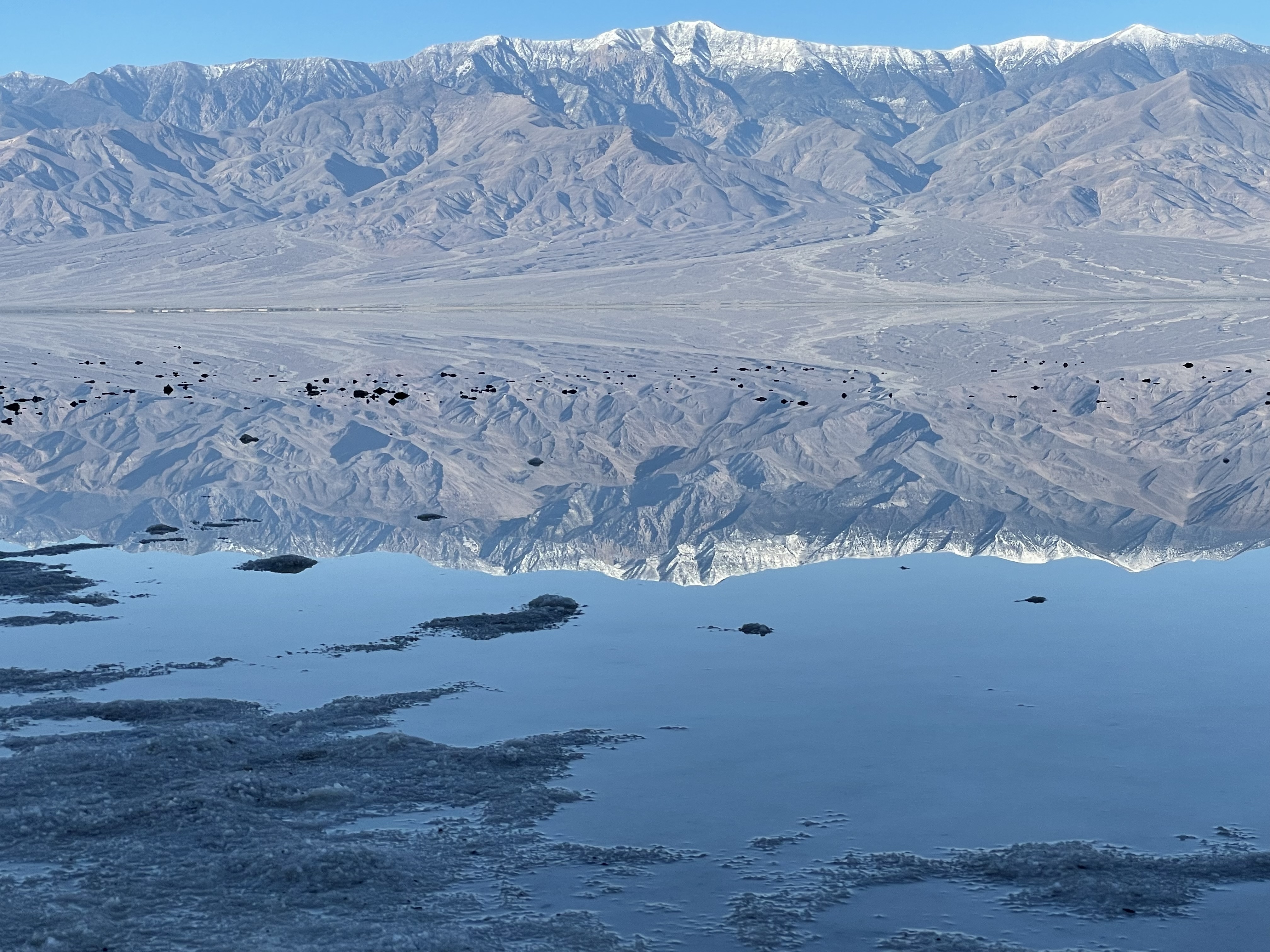 A snow-capped brown mountain is reflected upside-down in flat water.